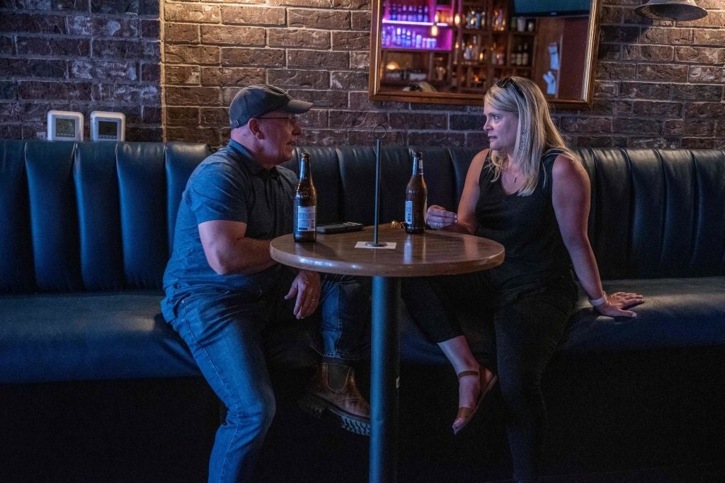 A man and a woman, neither wearing a mask, speak to each other at a bar table indoors
