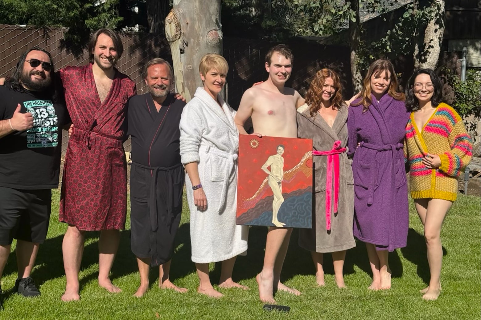 Seven actors smiling and wearing bathrobes outside. 