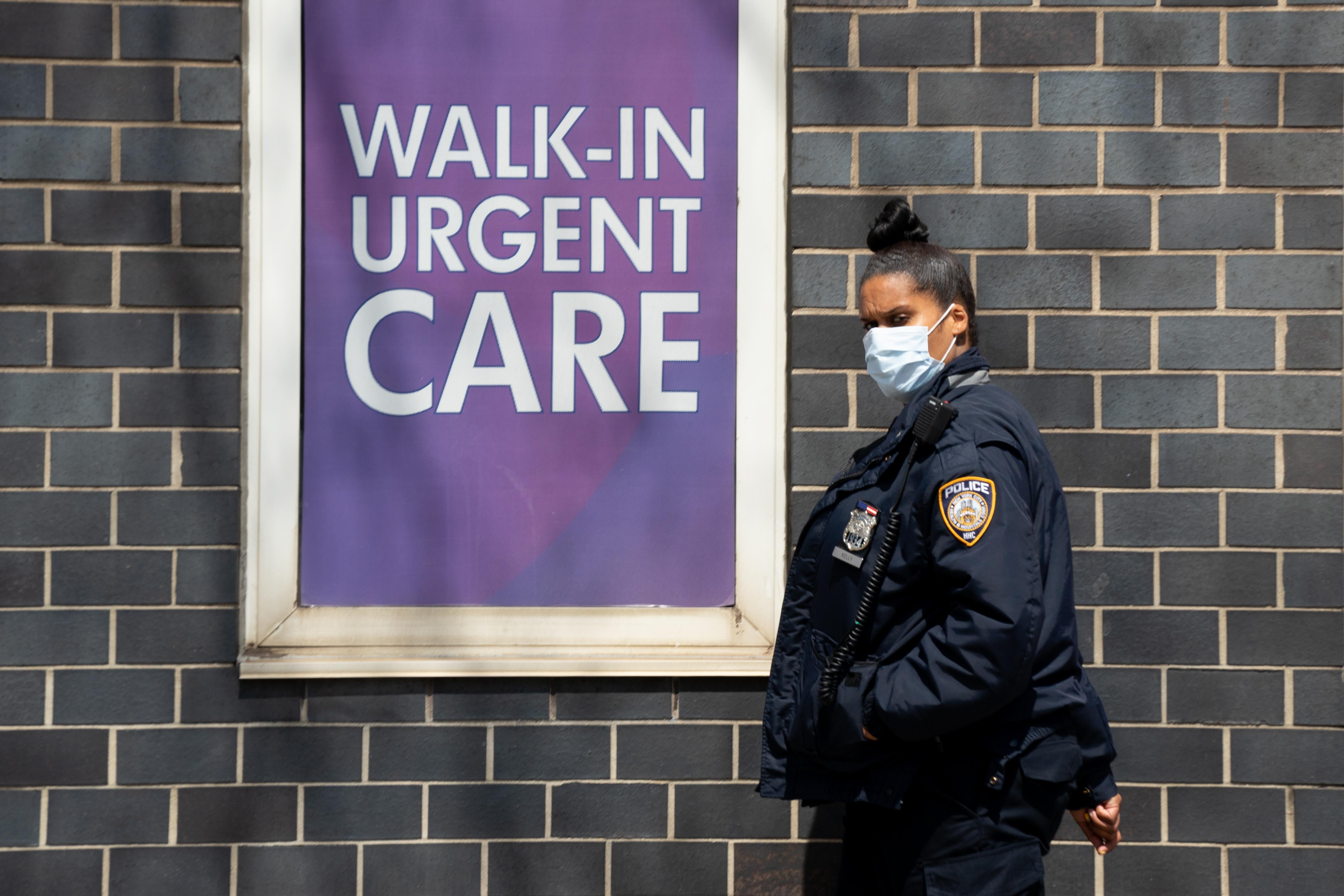 A police officer in a face mask walks near a sign that says "Walk-In Urgent Care."
