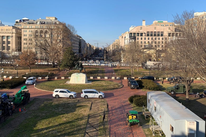 A park with several vehicles and trailers on it, a statue in the middle, and a fence at the back edge with a street stretching out into the distance.