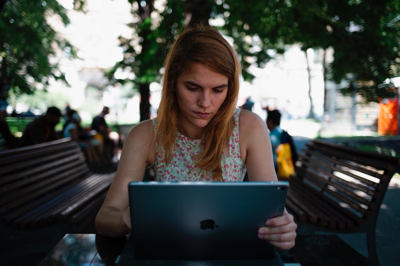 A woman sits at a table in a park, looking with consternation at her iPad screen.