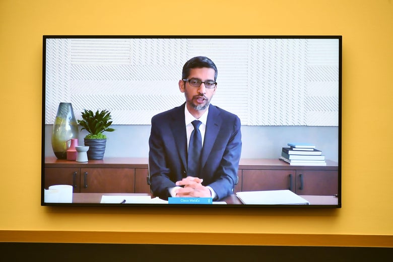 A screen on a yellow wall shows a man in a suit sitting at a desk, hands clasped.