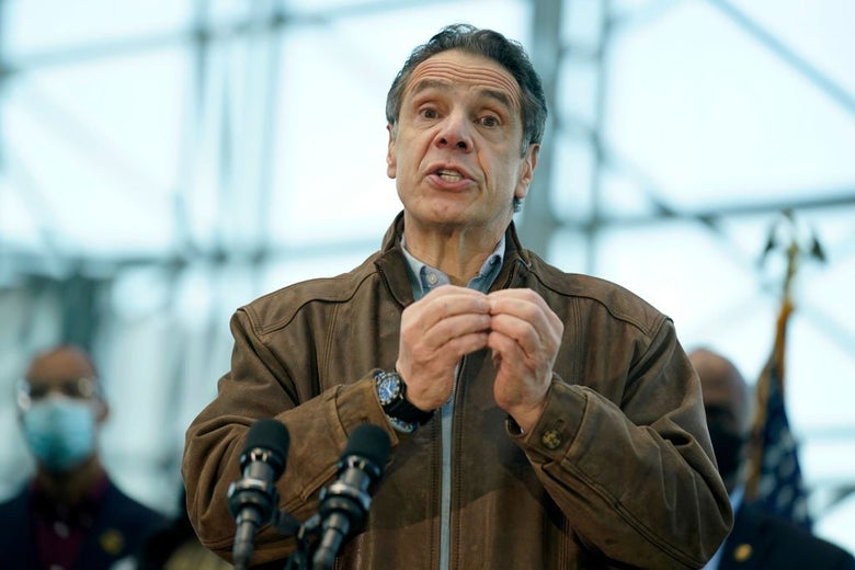 Cuomo, wearing a brown jacket, gestures while speaking into a microphone