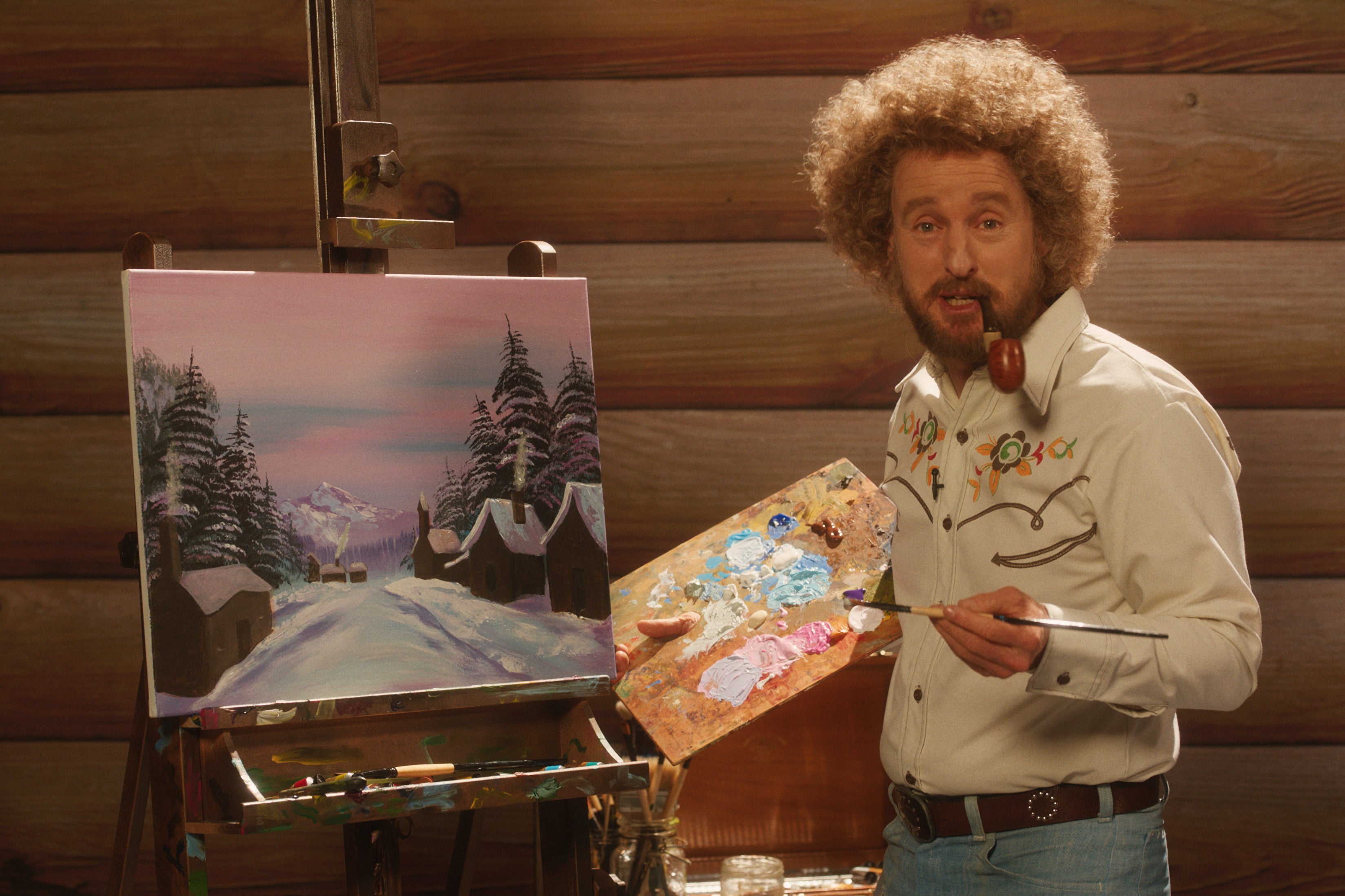 The actor stands in front of a painting of a wintry old-timey street lined with snow-capped pines, a palette in his right hand and a pipe in his mouth. He wars faded jeans, a Western shirt, and some very large permed hair.  