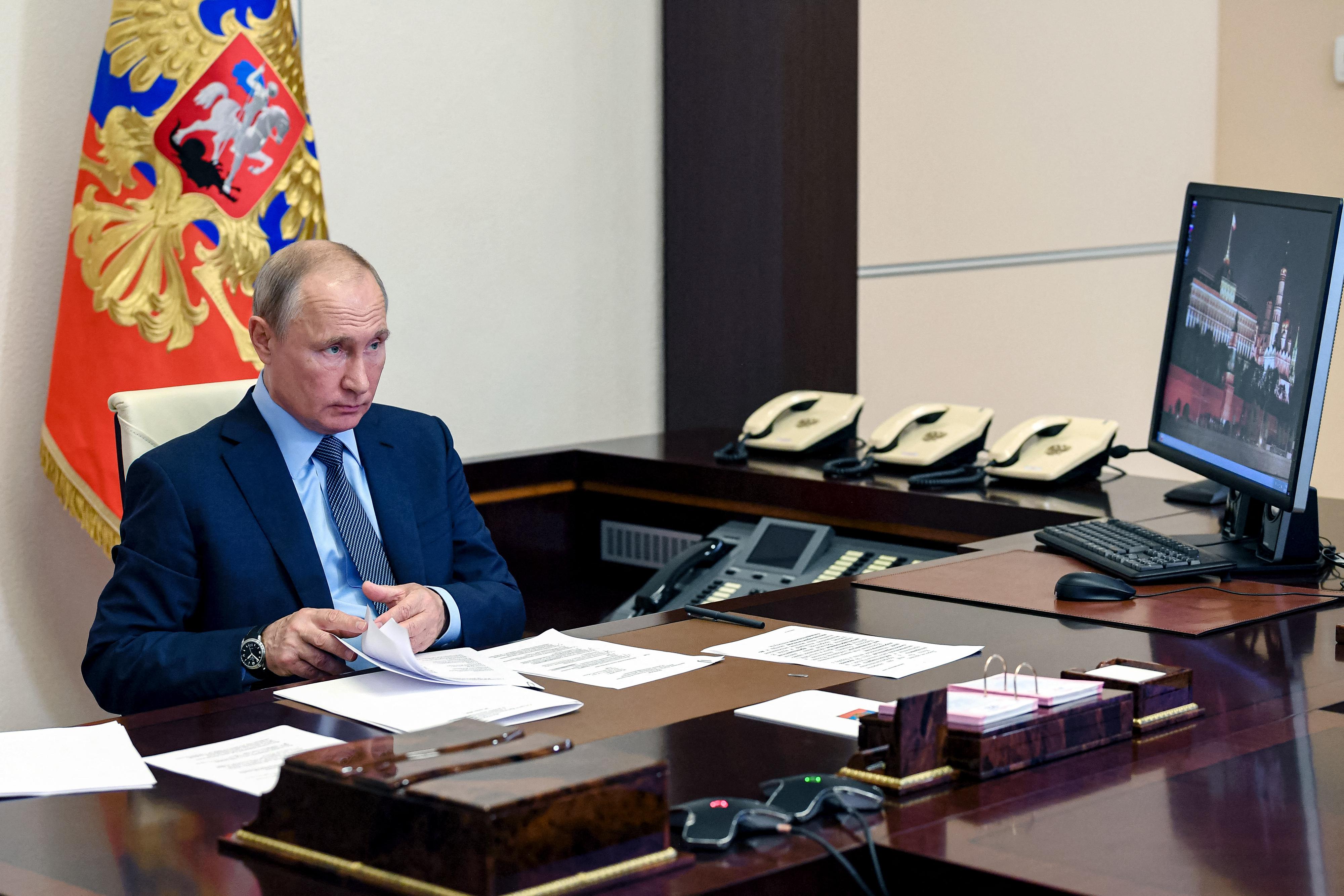 Putin sits at a desk with multiple phones and a large computer monitor in front of a flag.