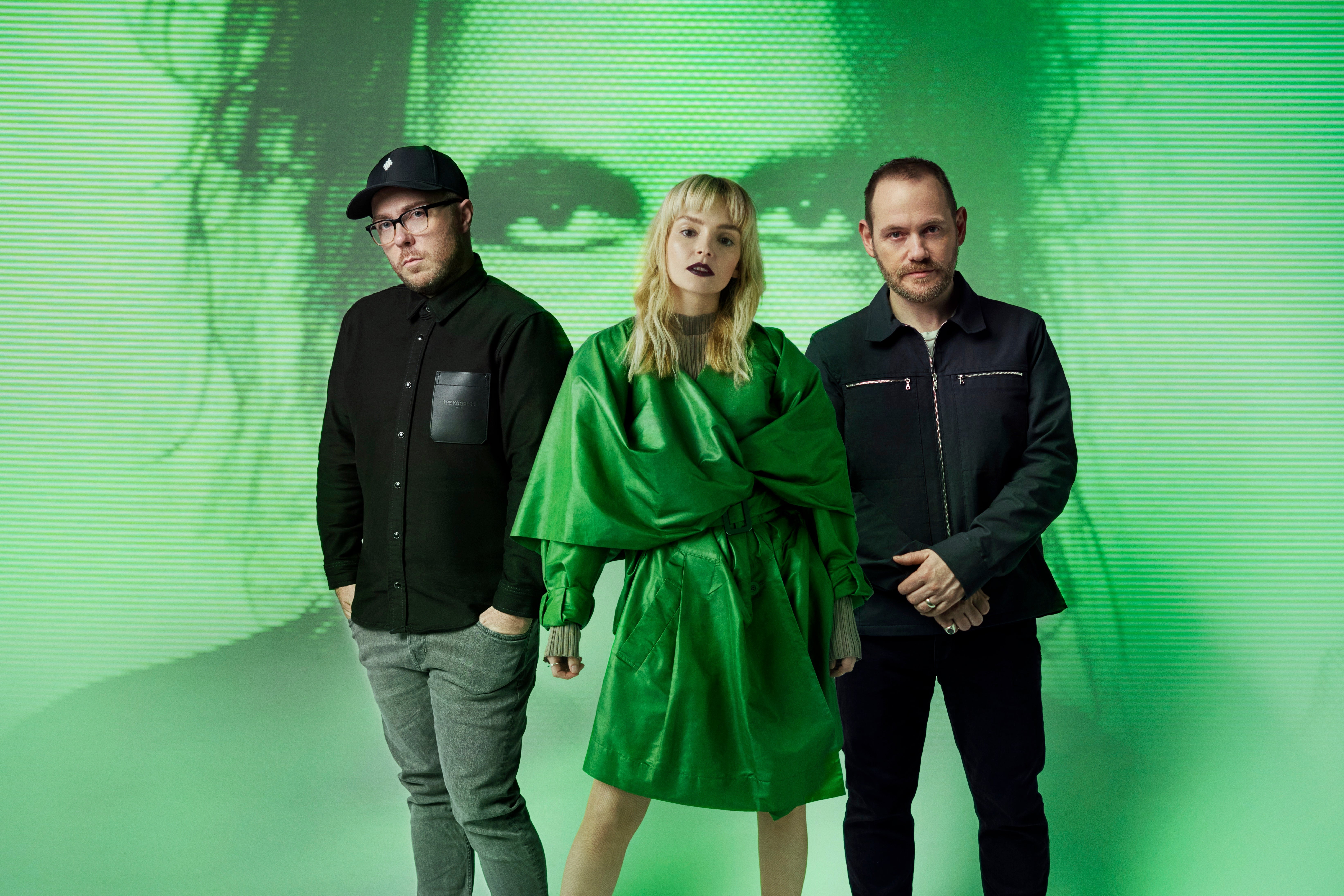 Three people—a man dressed in black with gray pants, a woman in a green dress with blonde hair, and a man in a navy jacket in black pants—stand in front of a green projection on the wall of a man holding his face. 