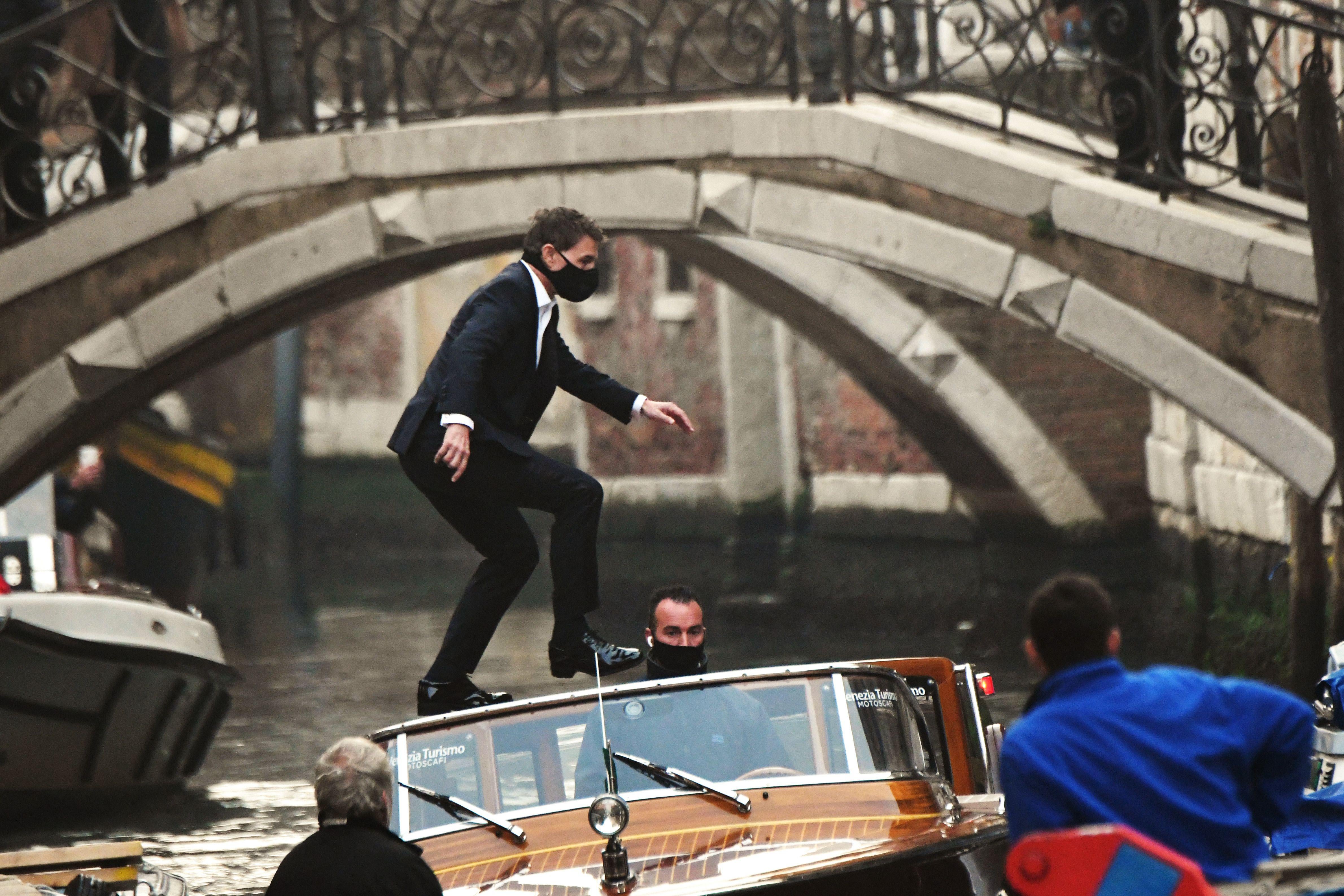 Tom Cruise, wearing a black mask, stands balancing on a taxi boat in a canal
