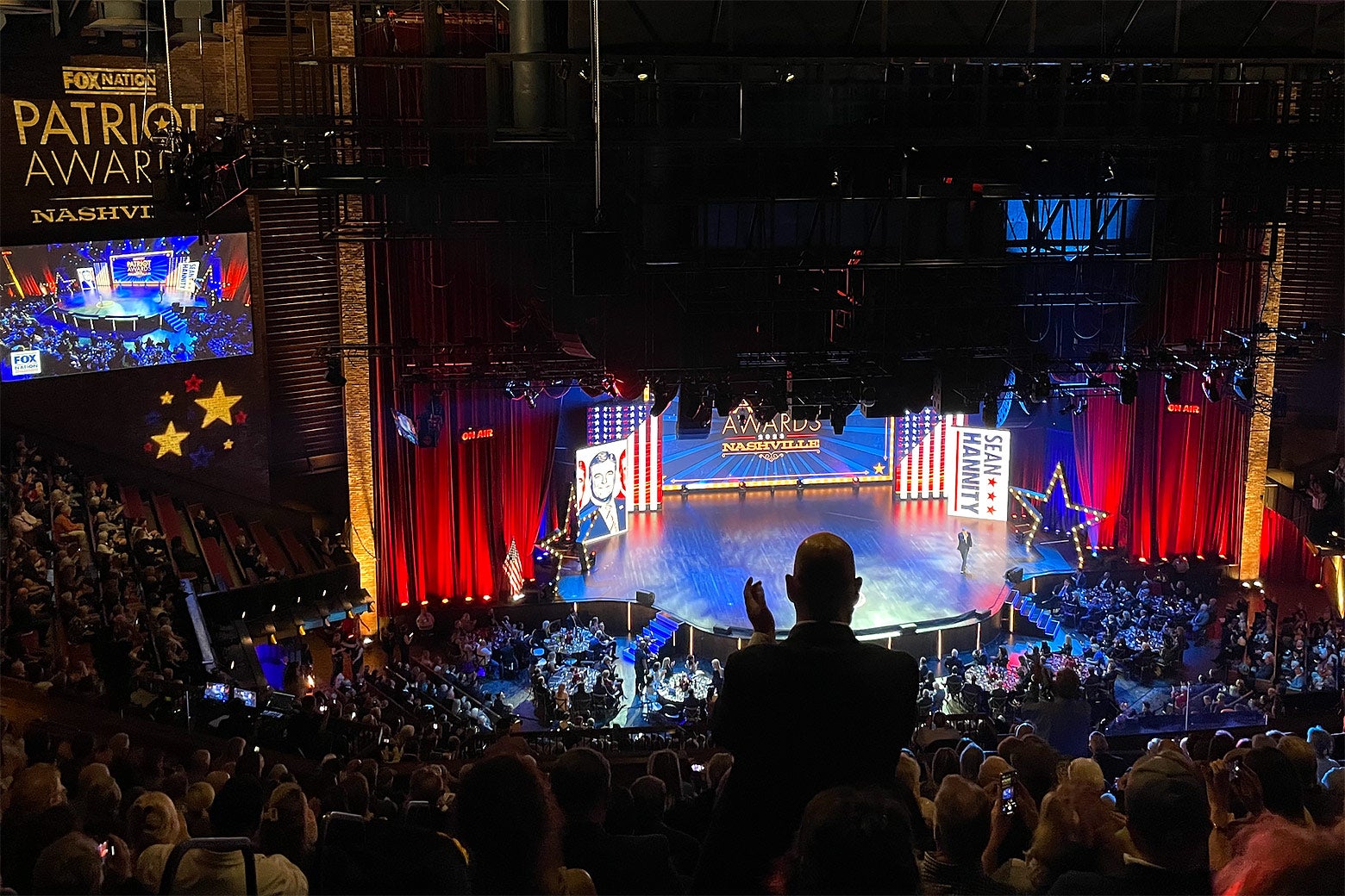 A man, in silhouette, stands to clap for Sean Hannity who is walking across a brightly lit stage at the Grand Ole Opry.