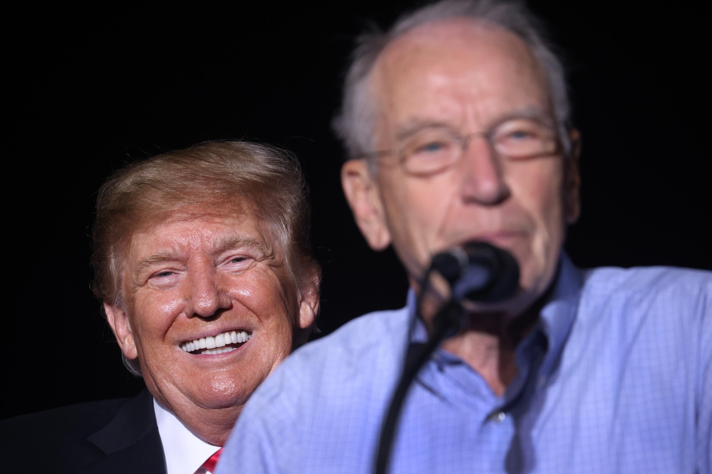 Trump in the background, smiling. Grassley in the foreground, speaking into a mic.