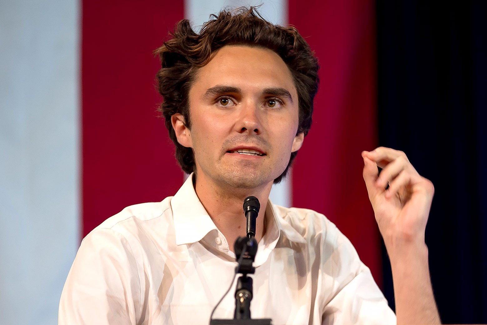 David Hogg is seated and relaxed in front of a microphone with his hand up gesturing.
