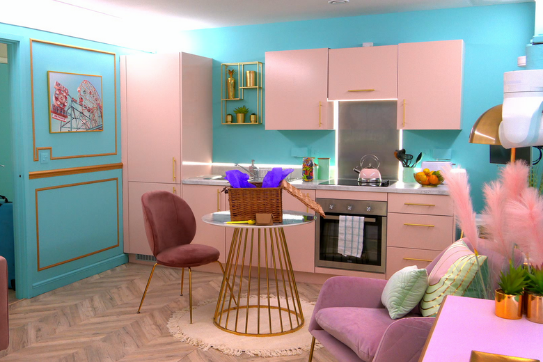 A room with turquoise blue walls and pink cabinets. There is a floral painting hanging on the wall behind a single red dining chair at a gold and white small table. In the corner is a dusty pink armchair with light green pillows. Next to it is a pink side table with two small plants sitting on top of it. 
