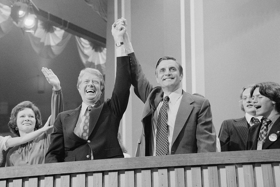 Carter and Mondale clasp hands at a stage while their families stand beside them.