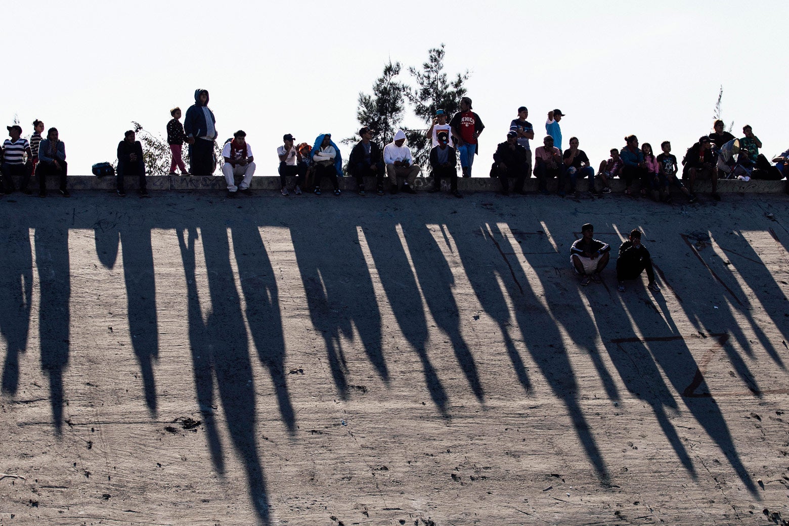 Migrants sitting on a concrete wall cast long shadows.