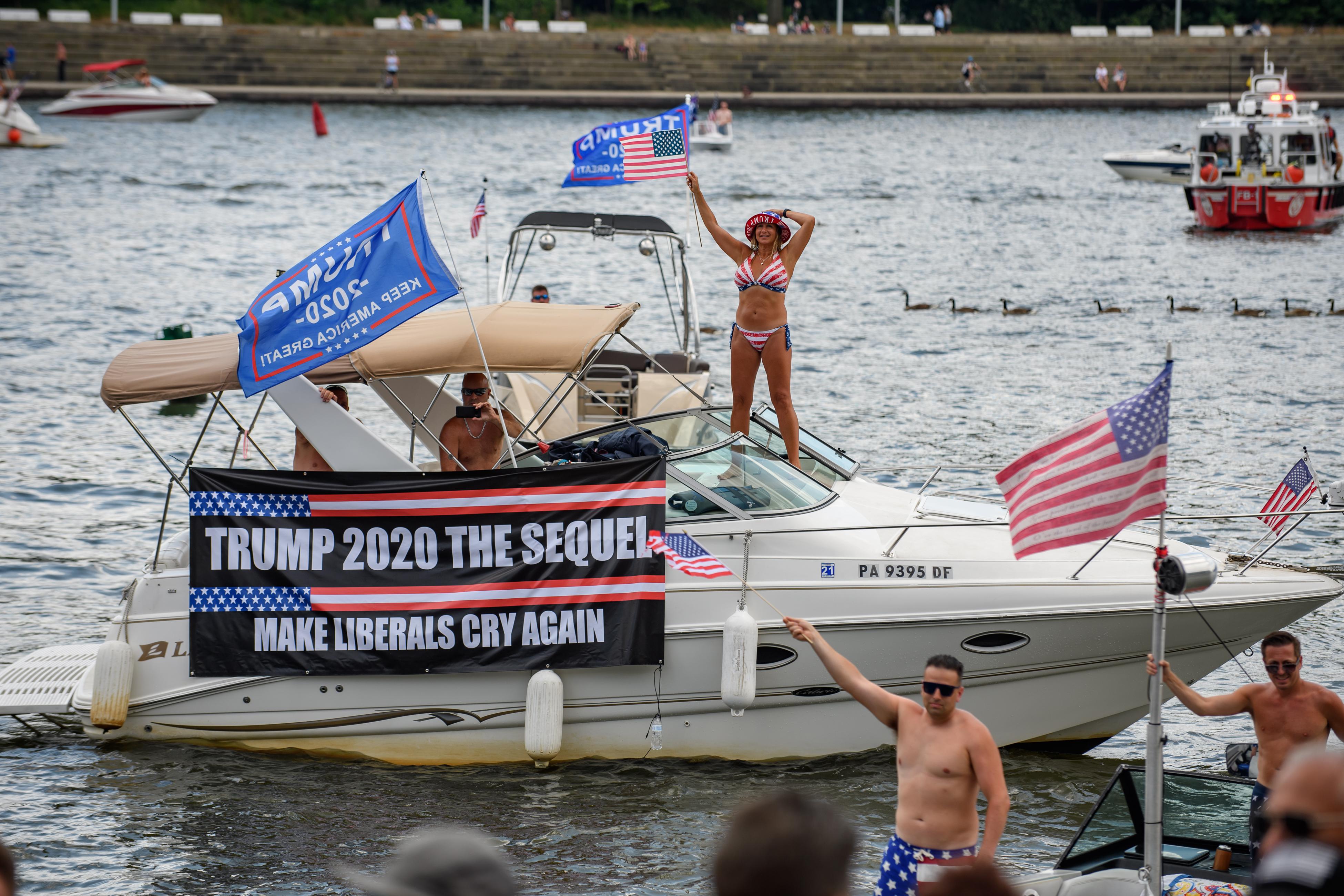 A yacht on a body of water. It has a pro-Trump banner hanging on it, a man driving holding up a Trump flag, and a woman standing on top of it in an American flag bikini, waving an American flag.