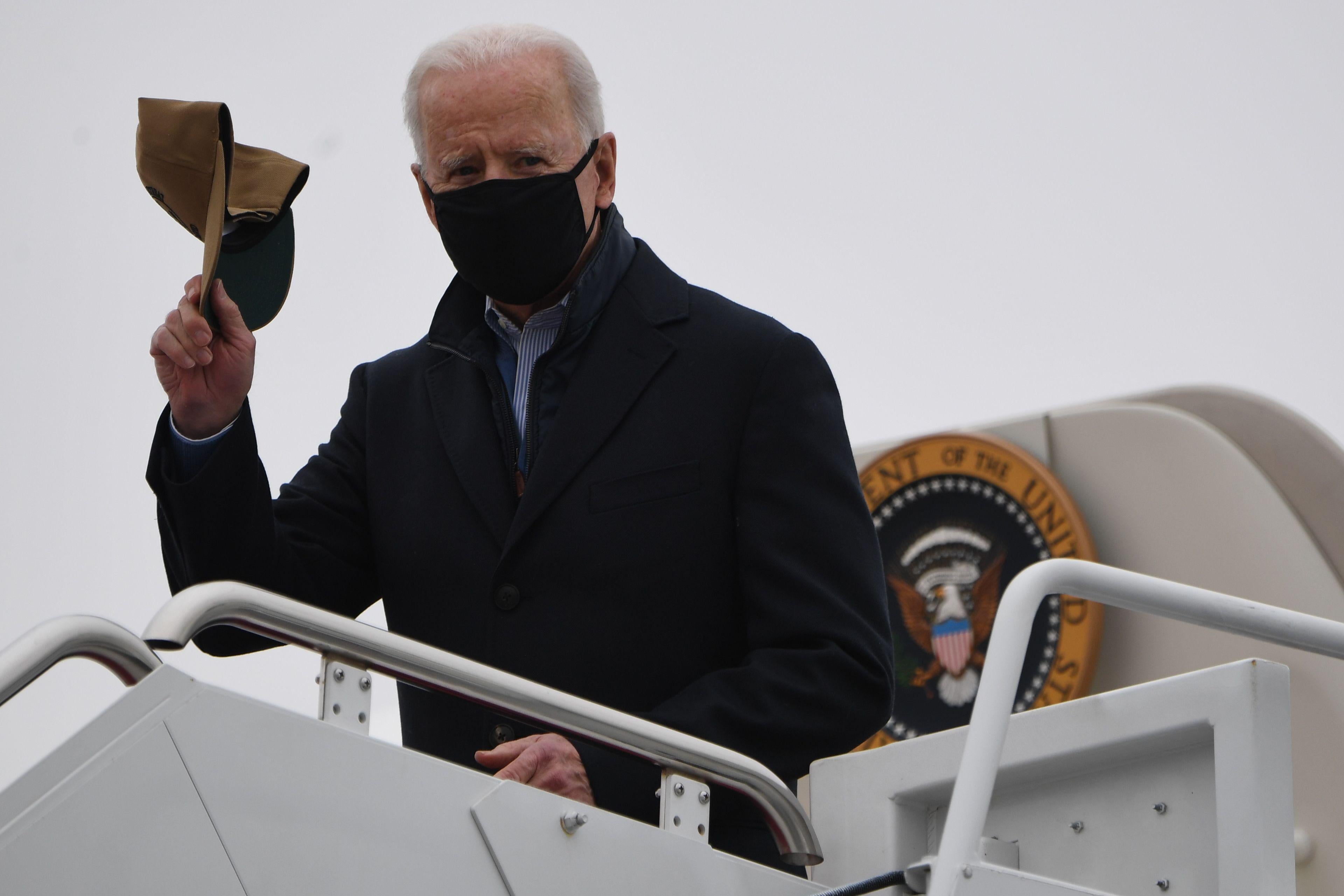 Biden stands at the top of the stairs disembarking from Air Force One, wearing a black mask and holding up a baseball cap