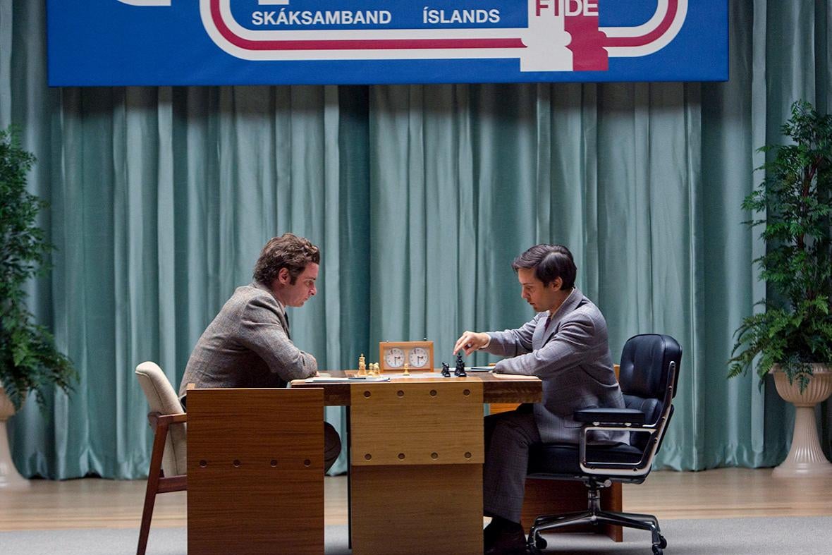 The two men sit at opposite sides of the chess board, a chess clock in the center