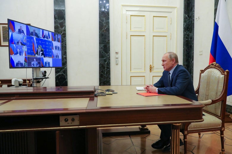 Putin sitting on the edge of his seat at a table in front of a monitor showing a video conference call