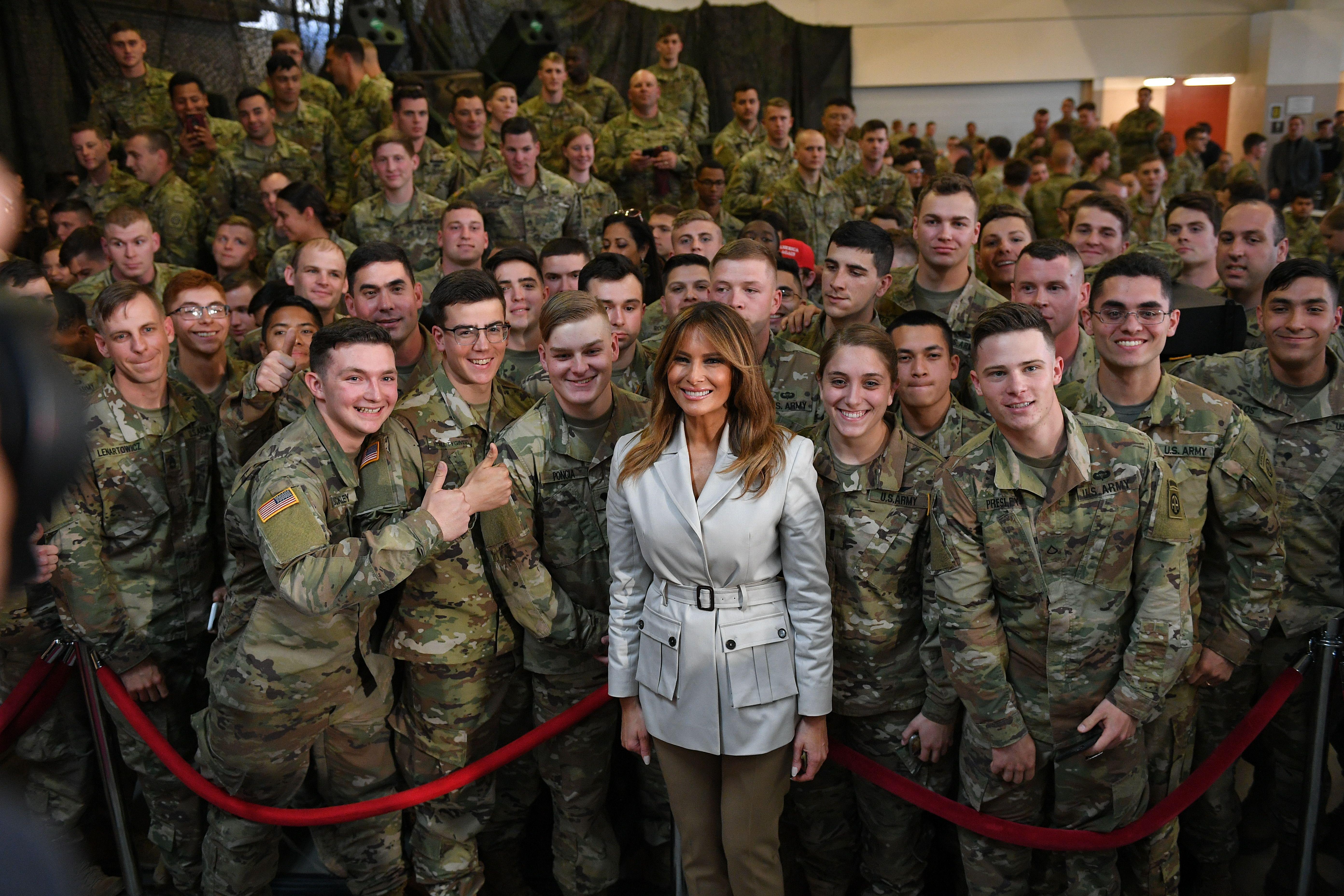 Melania Trump stands in front of a red velvet rope, behind which is a large group of people in camouflage military uniforms. Melania wears a belted taupe jacket with lapels and two cargo pockets, and slim olive green pants.