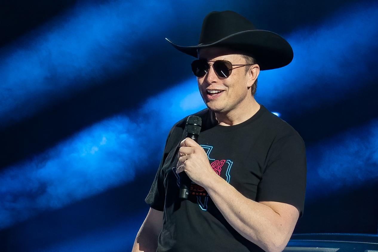 Elon Musk holds a mic while wearing sunglasses and a cowboy hat.
