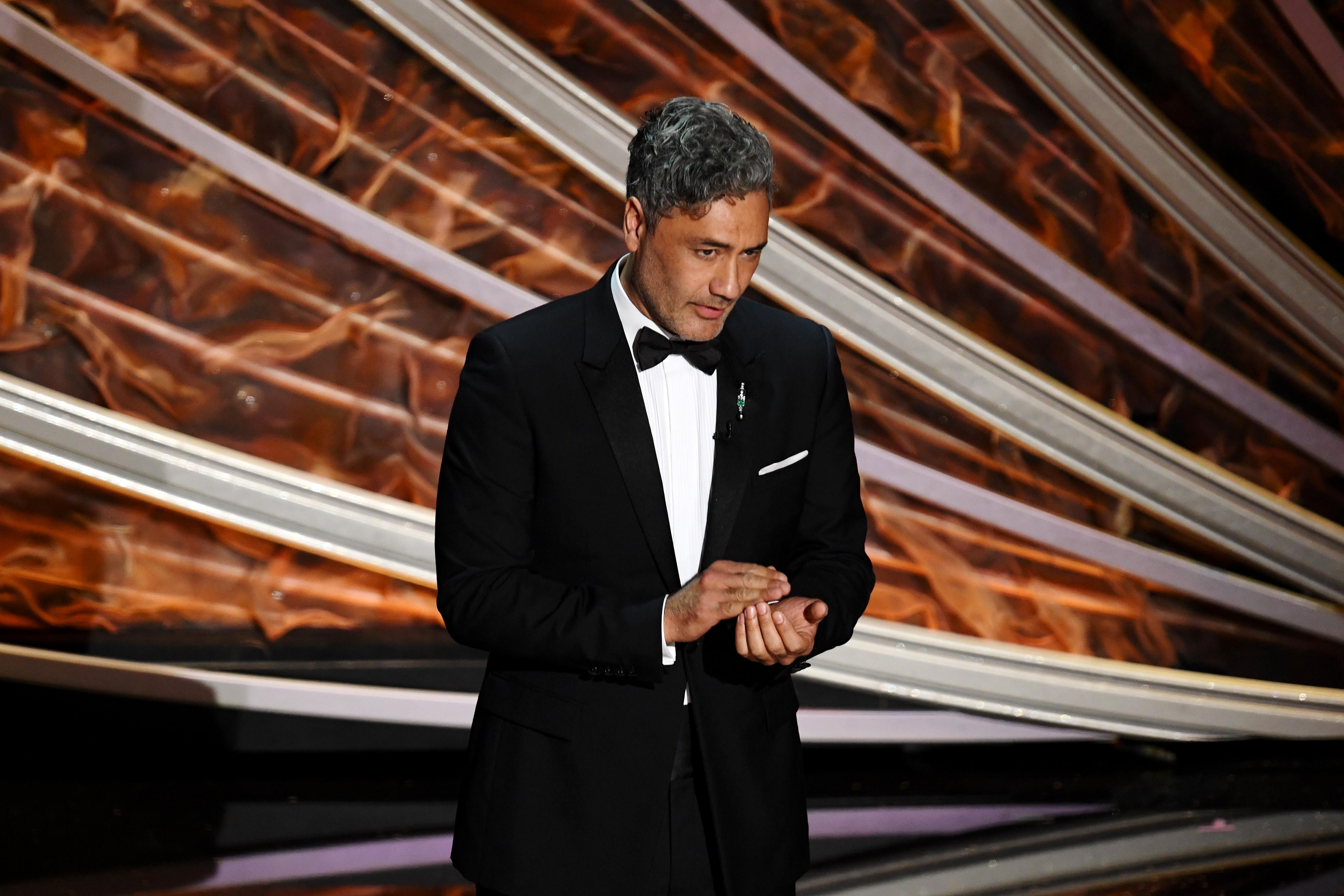 Taika Waititi, in a tux, speaking on a stage.