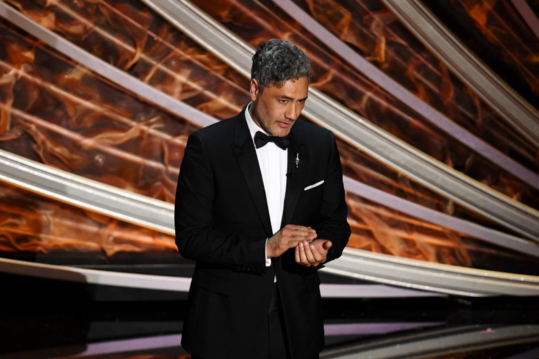 Taika Waititi, in a tux, speaking on a stage.