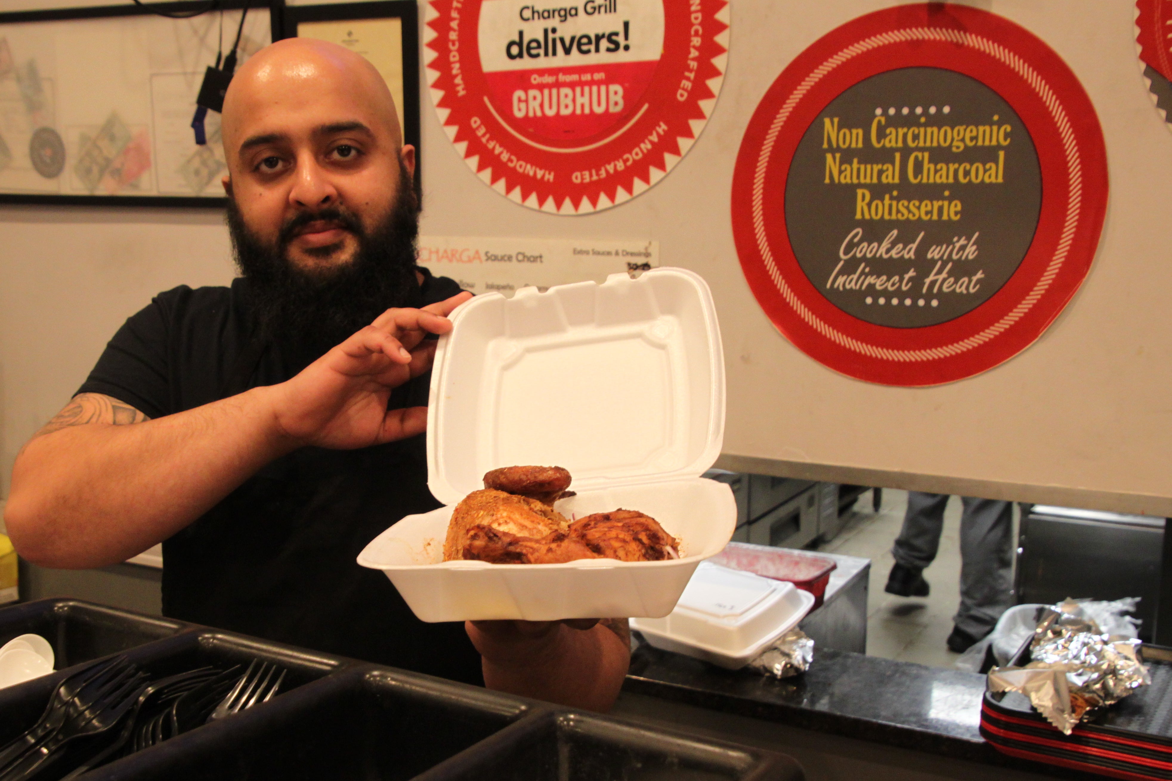 A man with a dark beard holds a Styrofoam container with a spiced roasted chicken inside.
