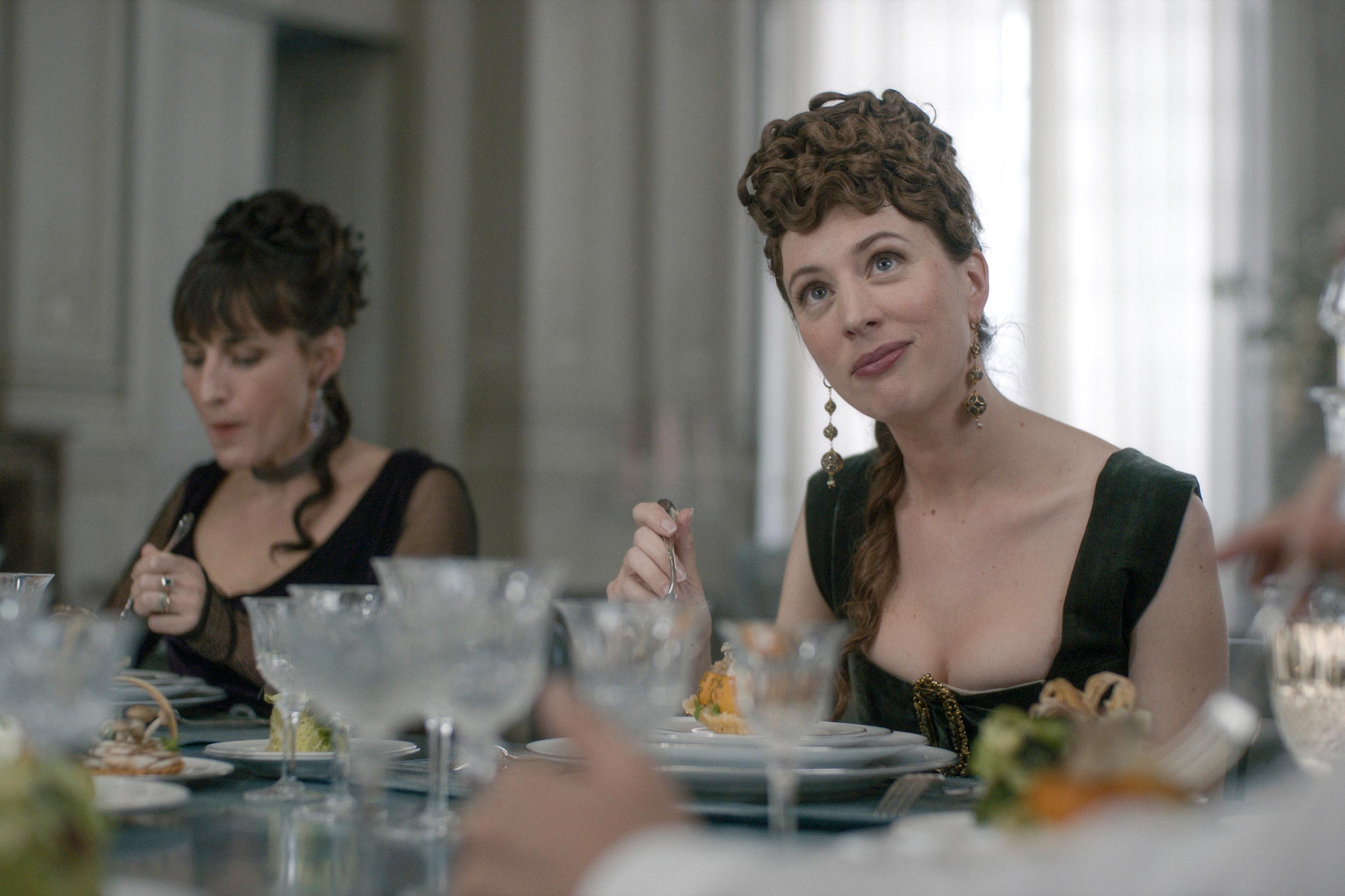 Two white women in nineteenth-century costumes eat fancy food at a table and look sly.