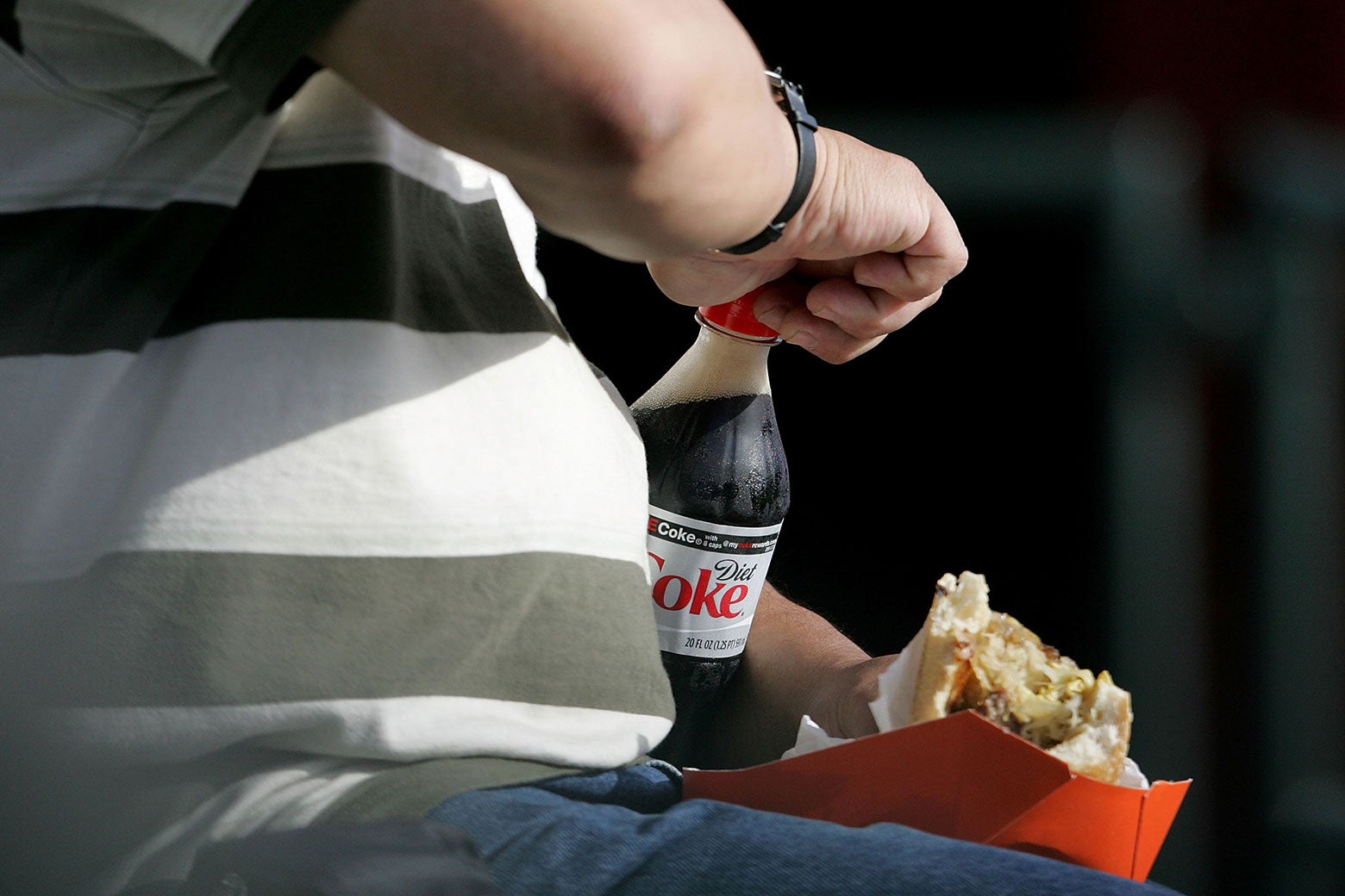 Un hombre abre una botella de Coca-Cola Light con una mano mientras sostiene un sándwich en la otra.