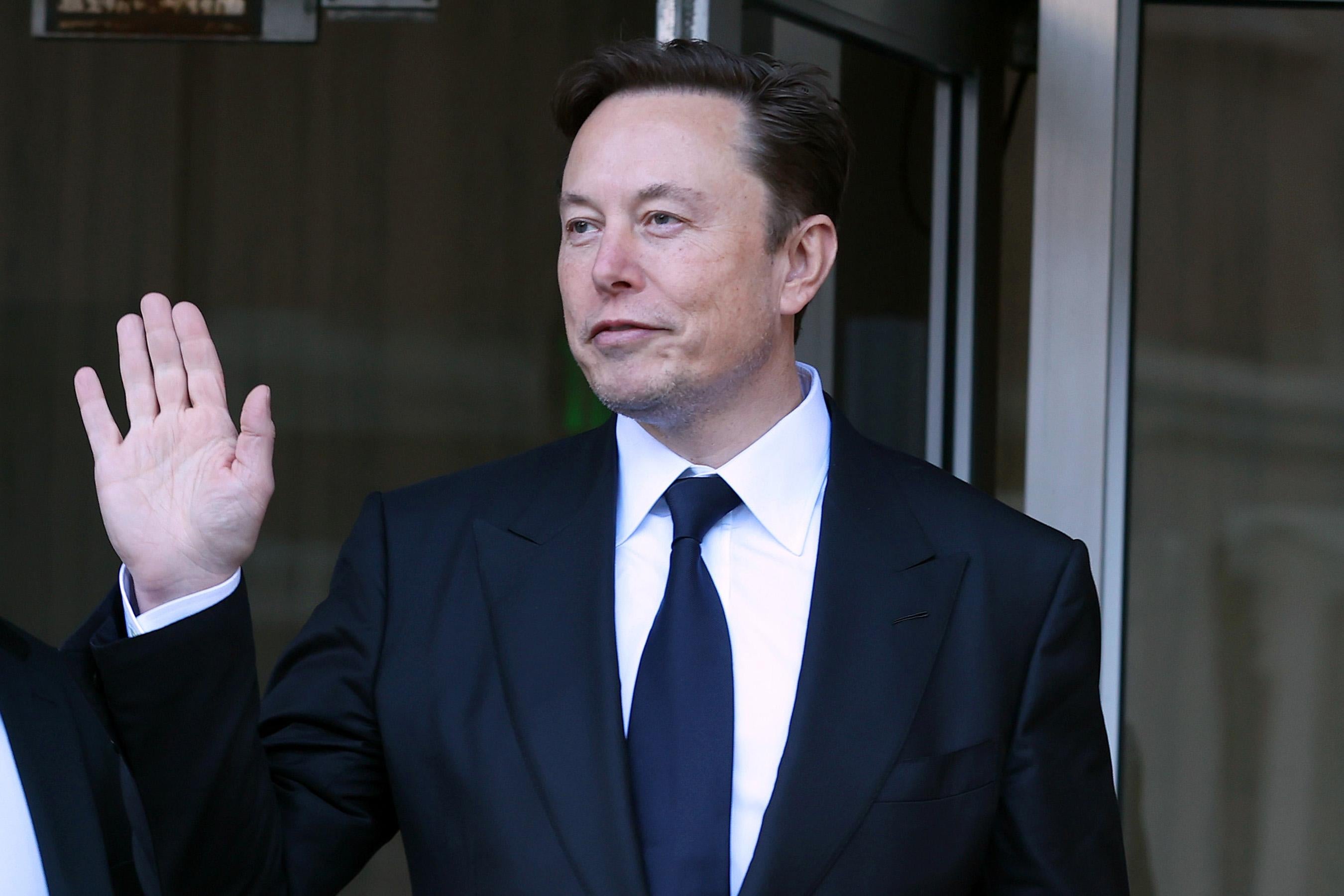 Elon Musk waves as he leaves a building wearing a suit and tie.