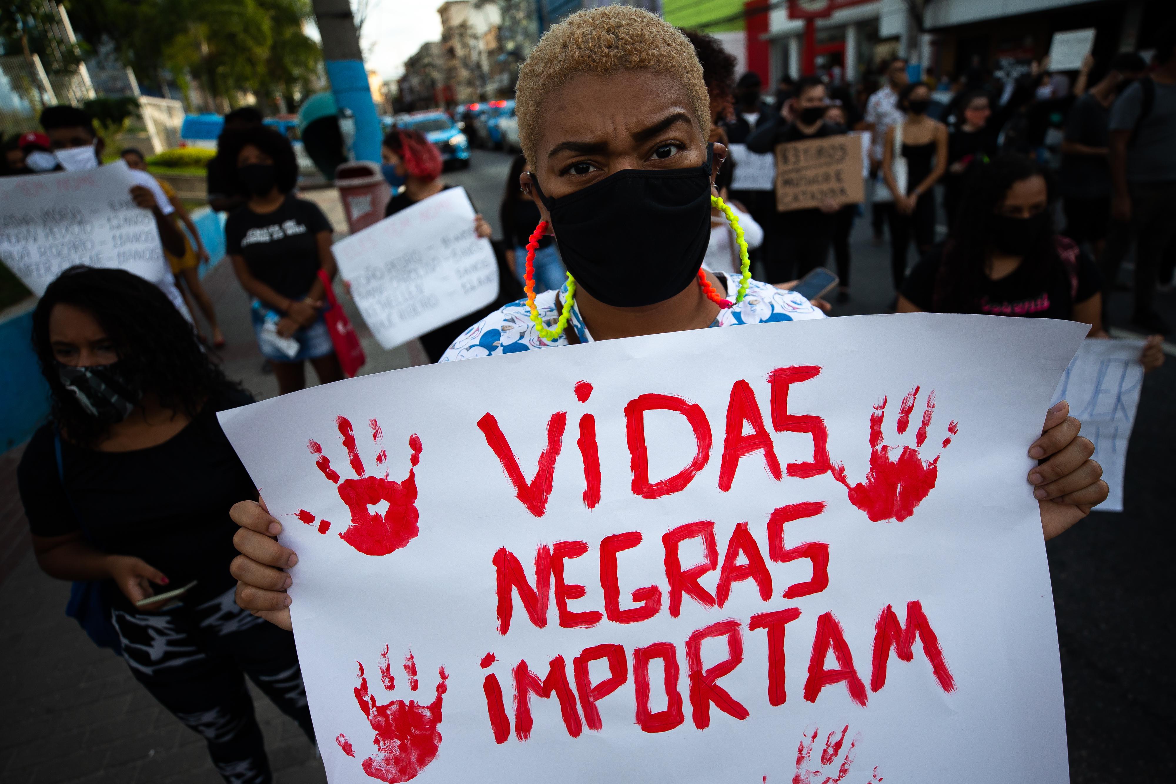A woman in a mask holds a red-painted sign reading "Black Lives Matter" in Portuguese at a rally.