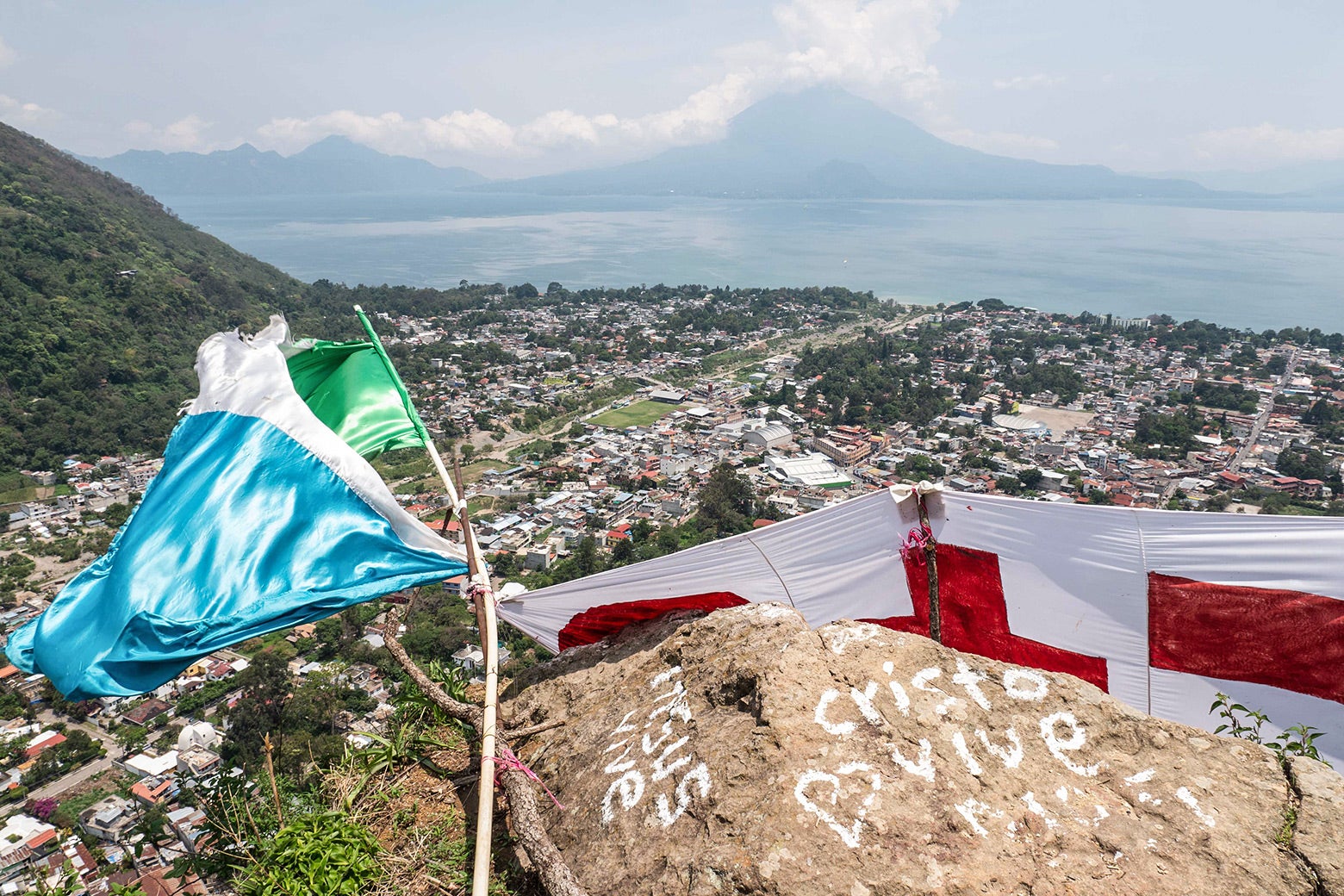 A view from above of a village nestled in mountains. Graffiti on a rock reads "Cristo Vive" next to a heart symbol.