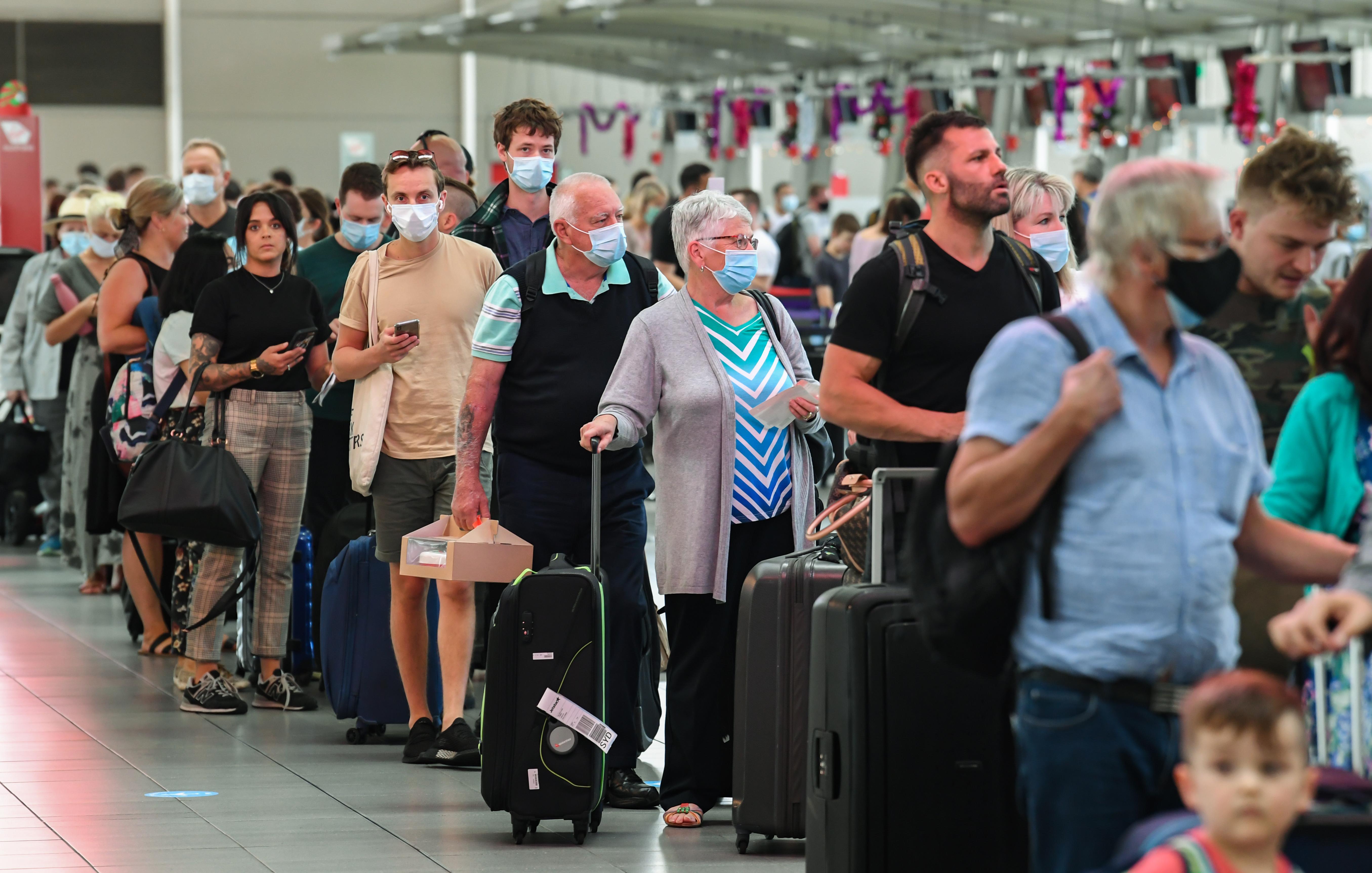 People, some wearing masks, stand in line with luggage in an airport.