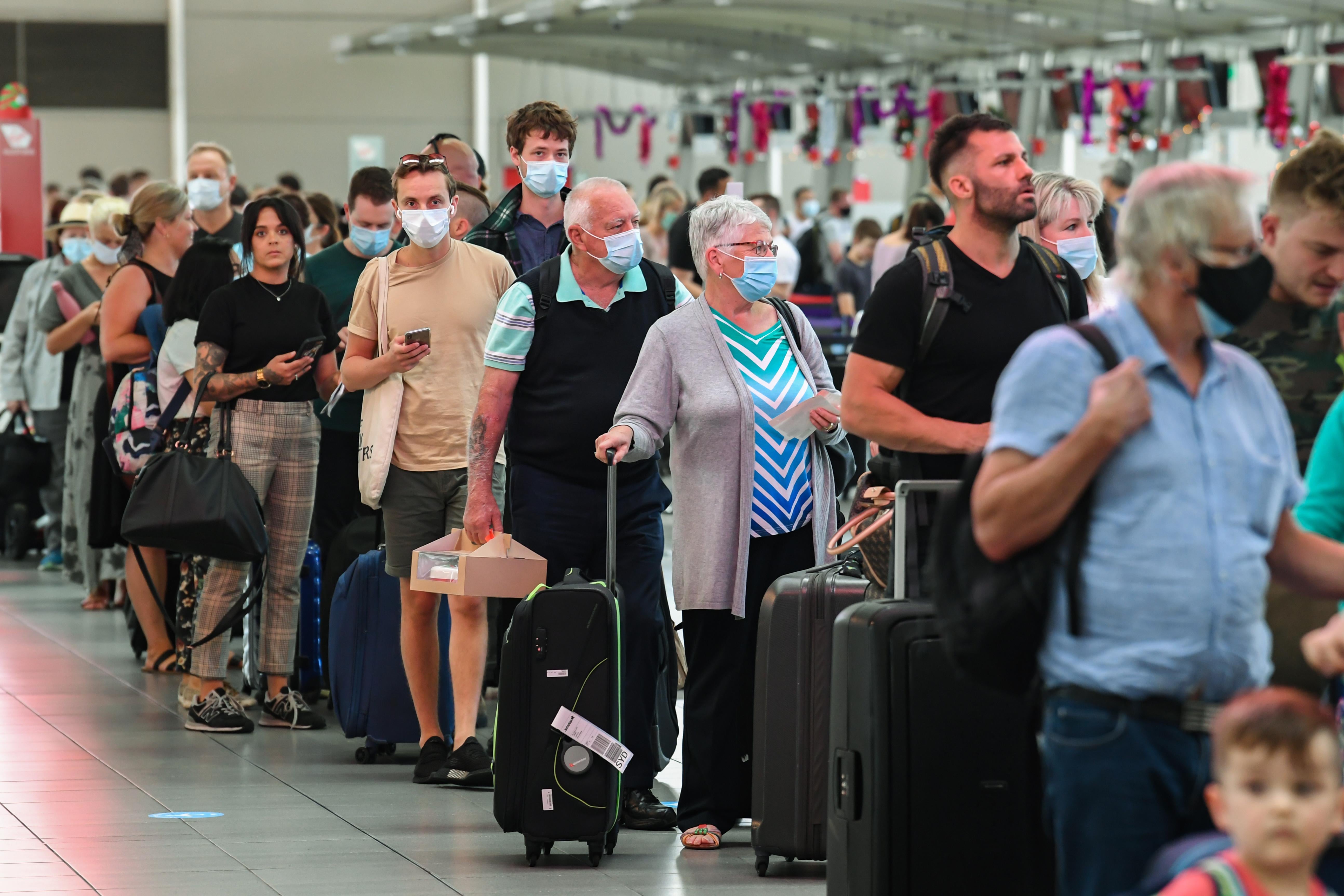 People, some wearing masks, stand in line with luggage in an airport.