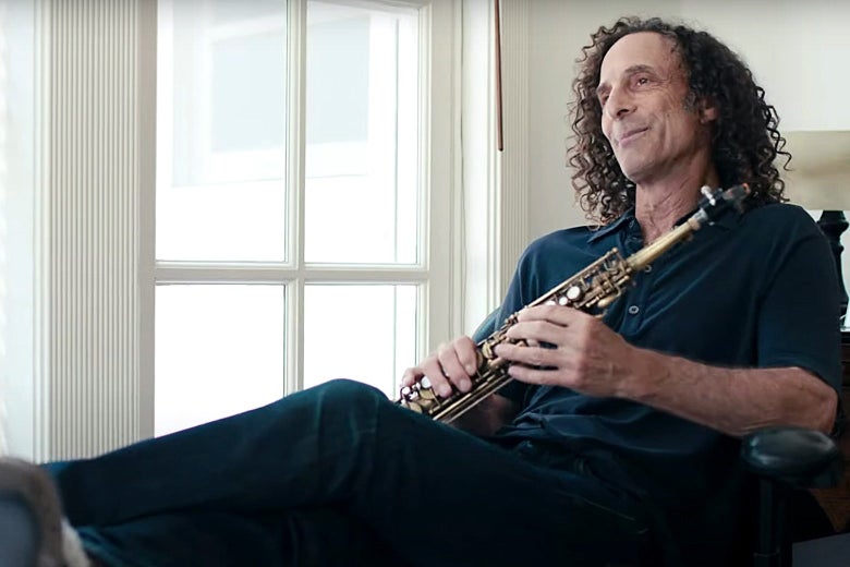 Kenny G reclined in a chair in fuzzy slippers while holding a soprano saxophone.