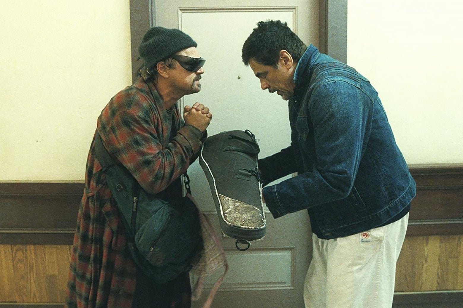 Two men talk in a hallway. One wears a checkered bathrobe and sunglasses, and the other, wearing a denim jacket, is holding a small package, likely a violin case, and attempting to hand it to the man in the robe.