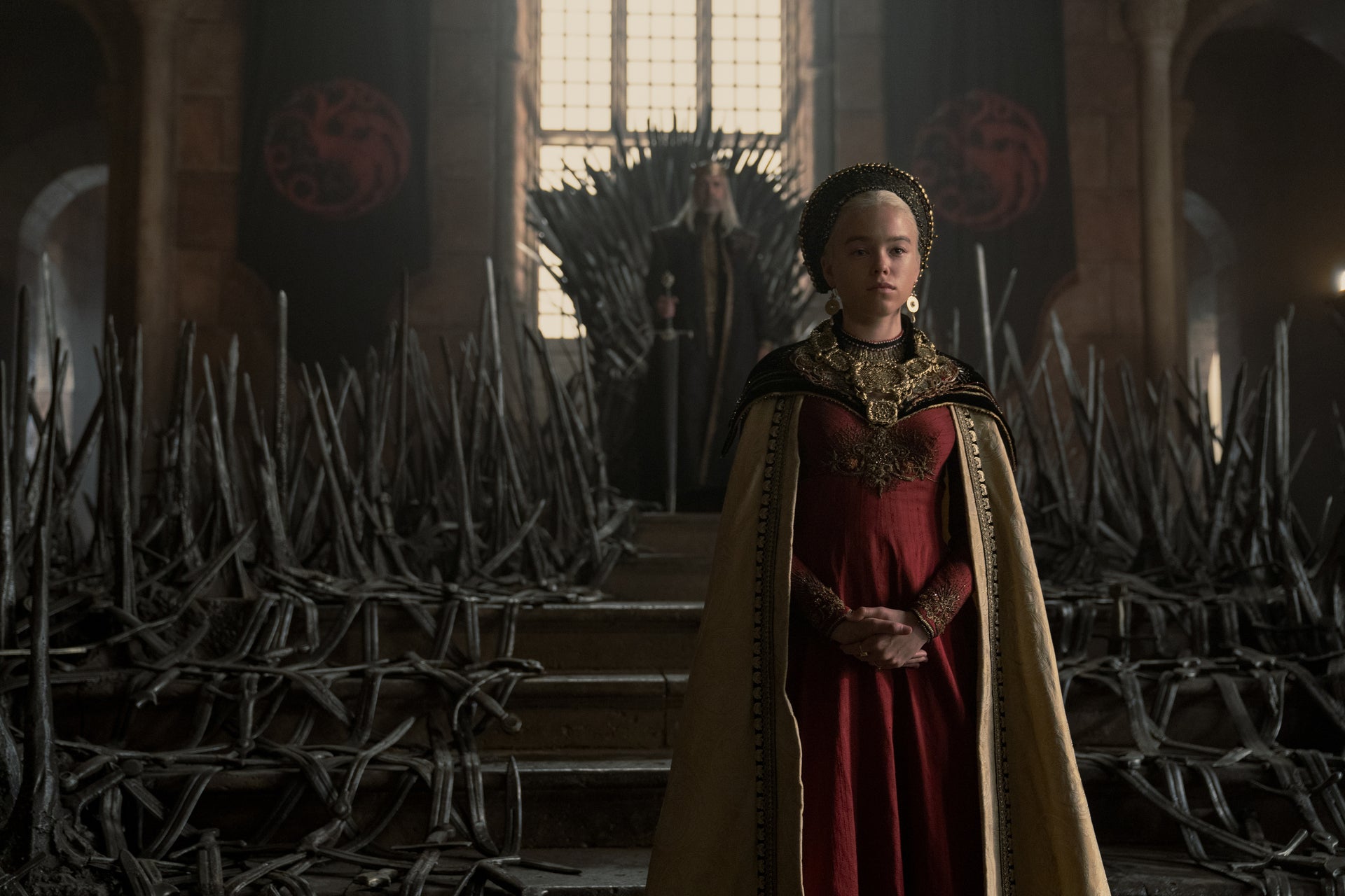 The young woman stands at the bottom of the steps below the familiar Iron Throne, facing away from the king, seated behind her. She wears a gold cloak, a red dress, and a determined expression.