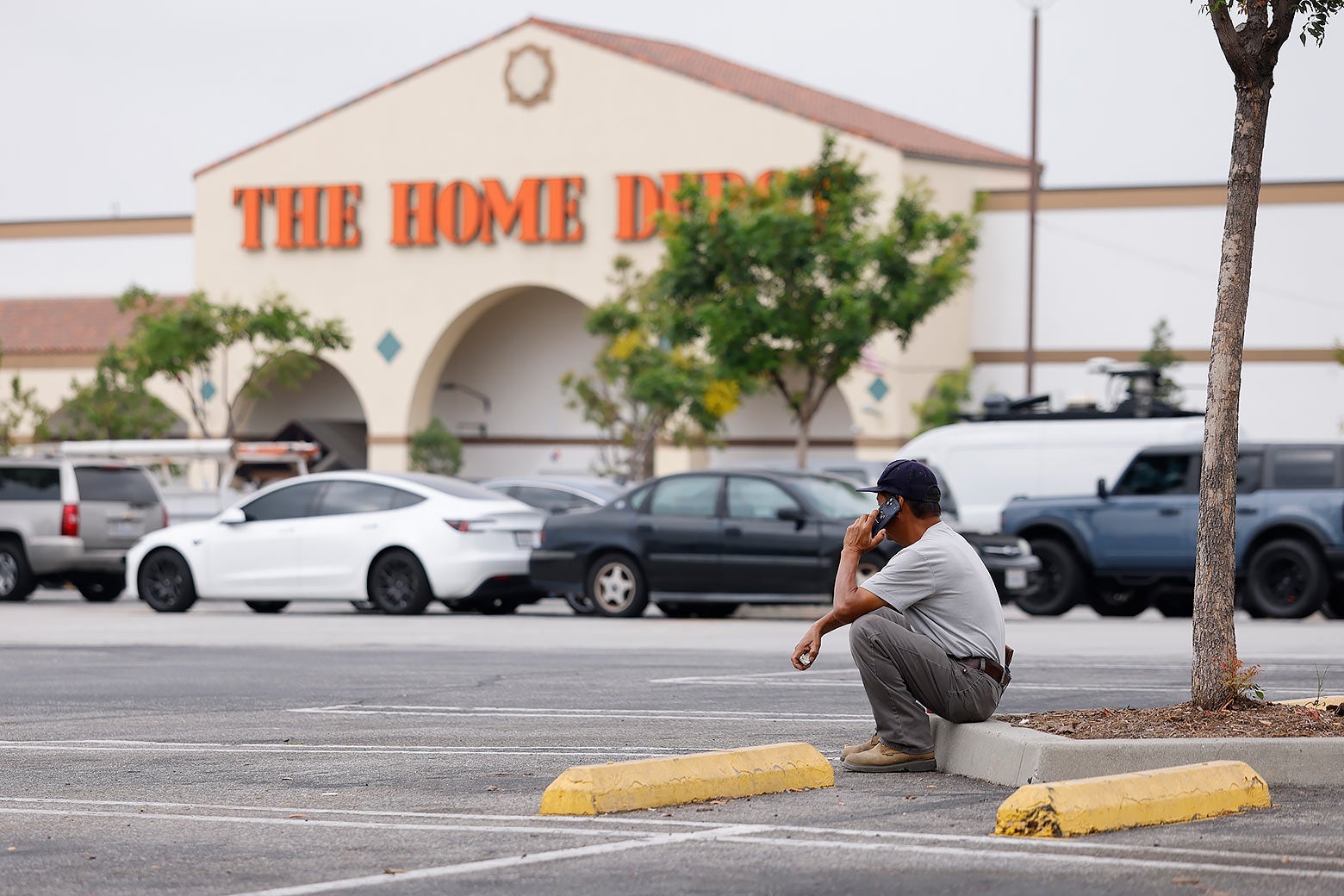 A lone man squats in a Home Depot parking lot talking on his cellphone.