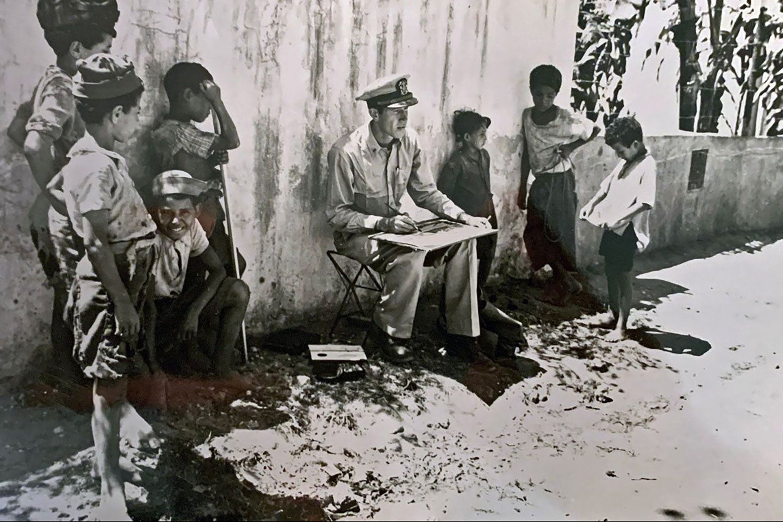 A man in military uniform sits with his back to a wall and is surrounded by standing children who look curious.