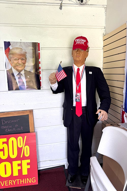 A full-sized mannequin modeled after Donald Trump in a suit, red tie, and a red hat reading "TRUMP 2028." A yellow "50% OFF EVERYTHING" sign stands to its left.