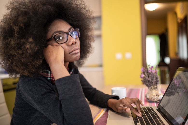 A Black woman in glasses rests her chin on her hand as she looks up from her laptop.