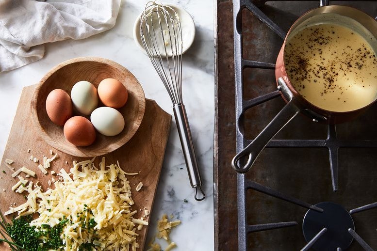 A bowl of five eggs and a mound of grated cheese and minced chives. Nearby, a pan filled with yellow sauce, on a stovetop.
