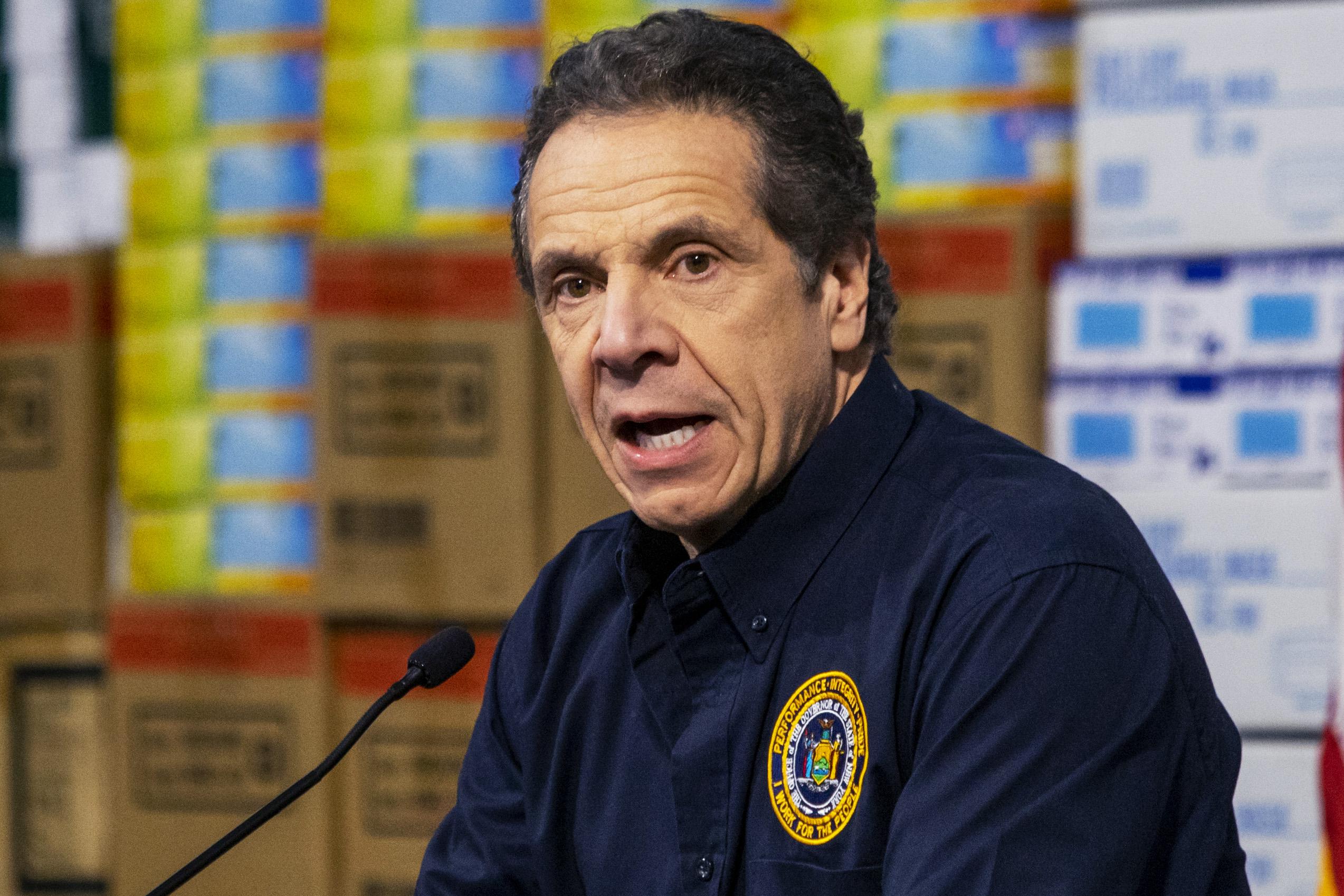 Cuomo speaks at a mic with boxes of supplies behind him