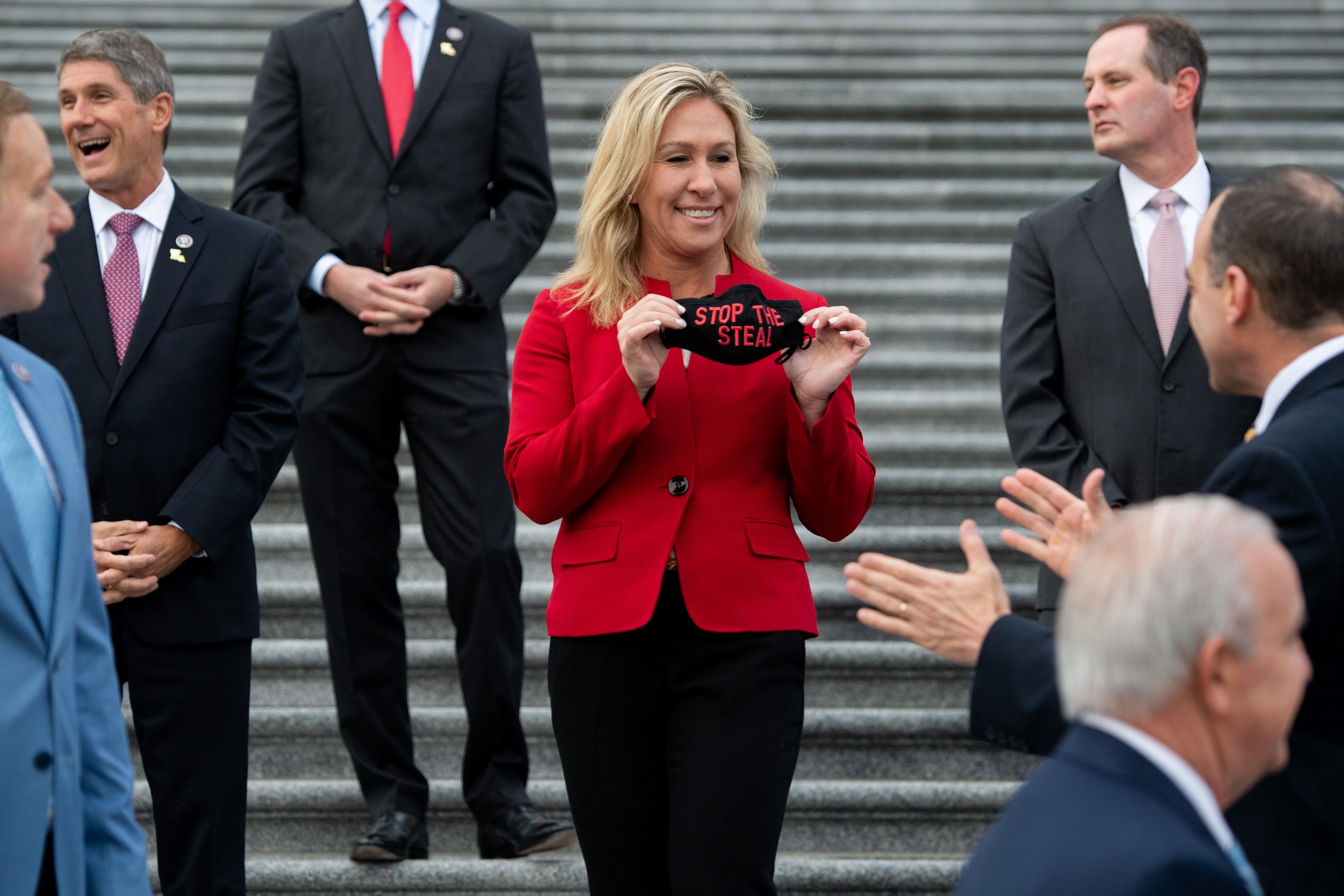Greene smiles as she holds up a mask that says "Stop the Steal" to supportive fellow Republican freshmen standing around her on the Capitol steps.