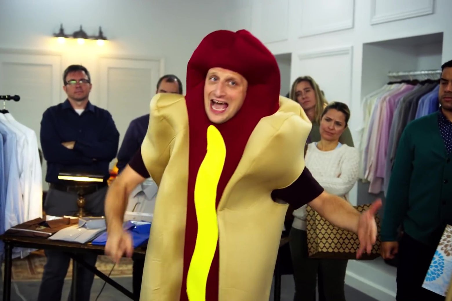 A man in a hot-dog costume stands amid a group of accusing people.
