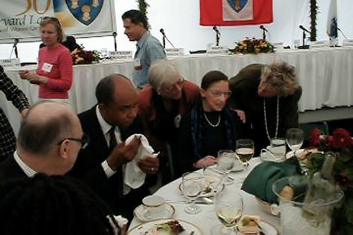 RBG at a table at an event with two women standing on either side of her to speak with her.