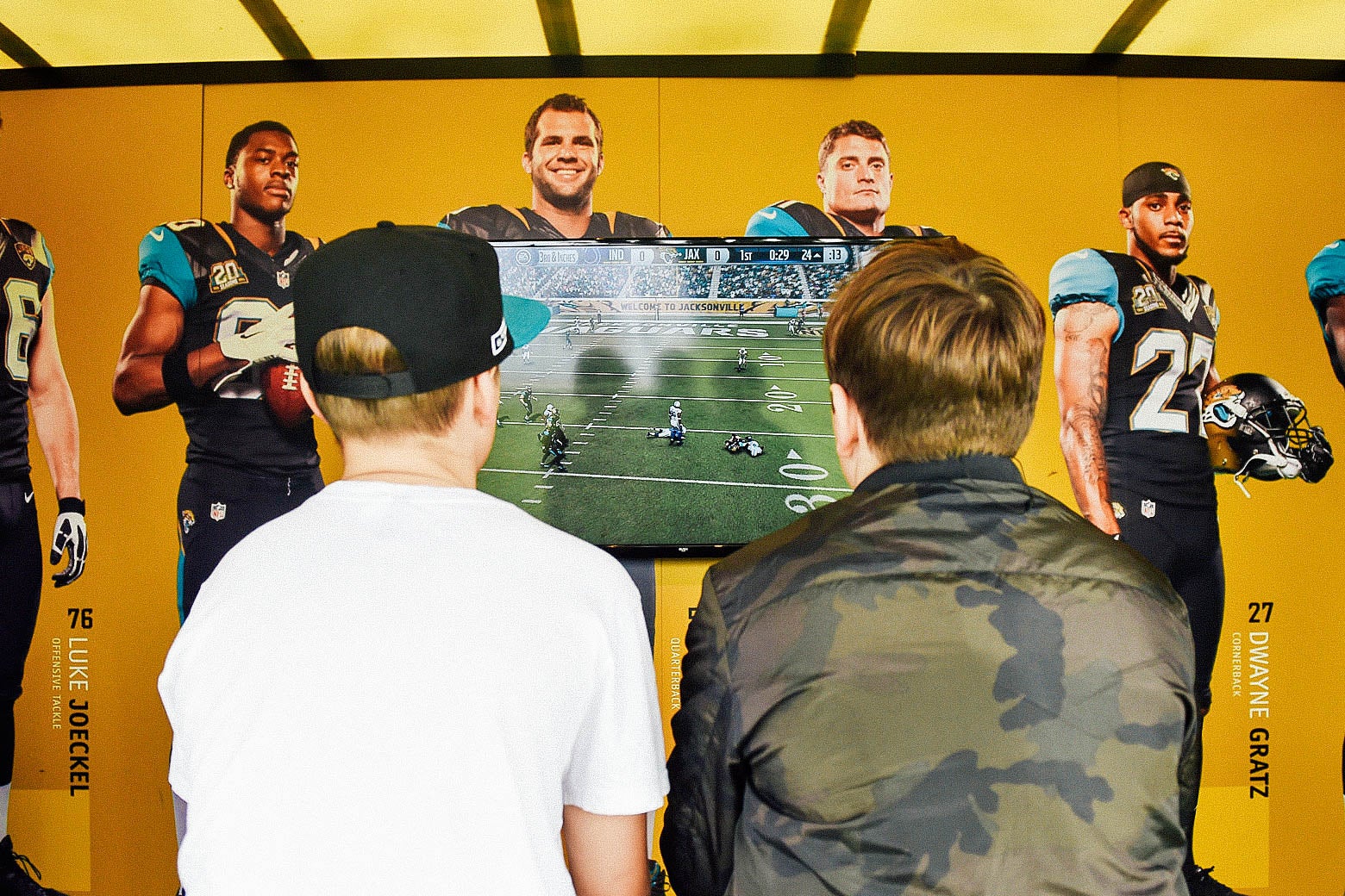 Two young men's backs are toward the gamer as they play Madden on a TV screen.