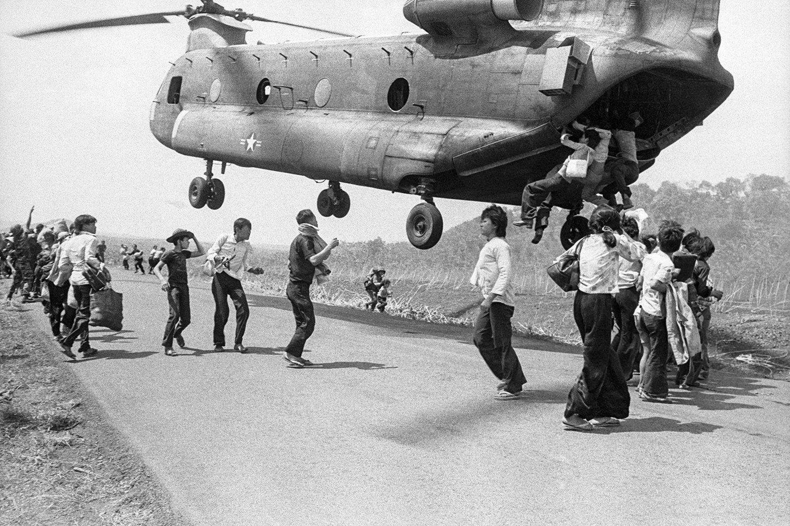 Vietnamese refugees stand on a runway as a U.S. helicopter hovers overhead with three people hanging from the back