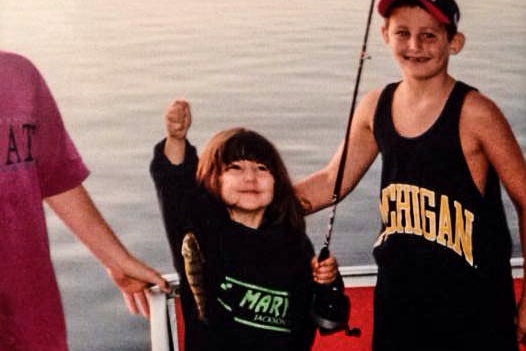 In the center, a young girl proudly holds a fishing reel and a small fish. Next to her is a grinning boy in a Michigan tank top.
