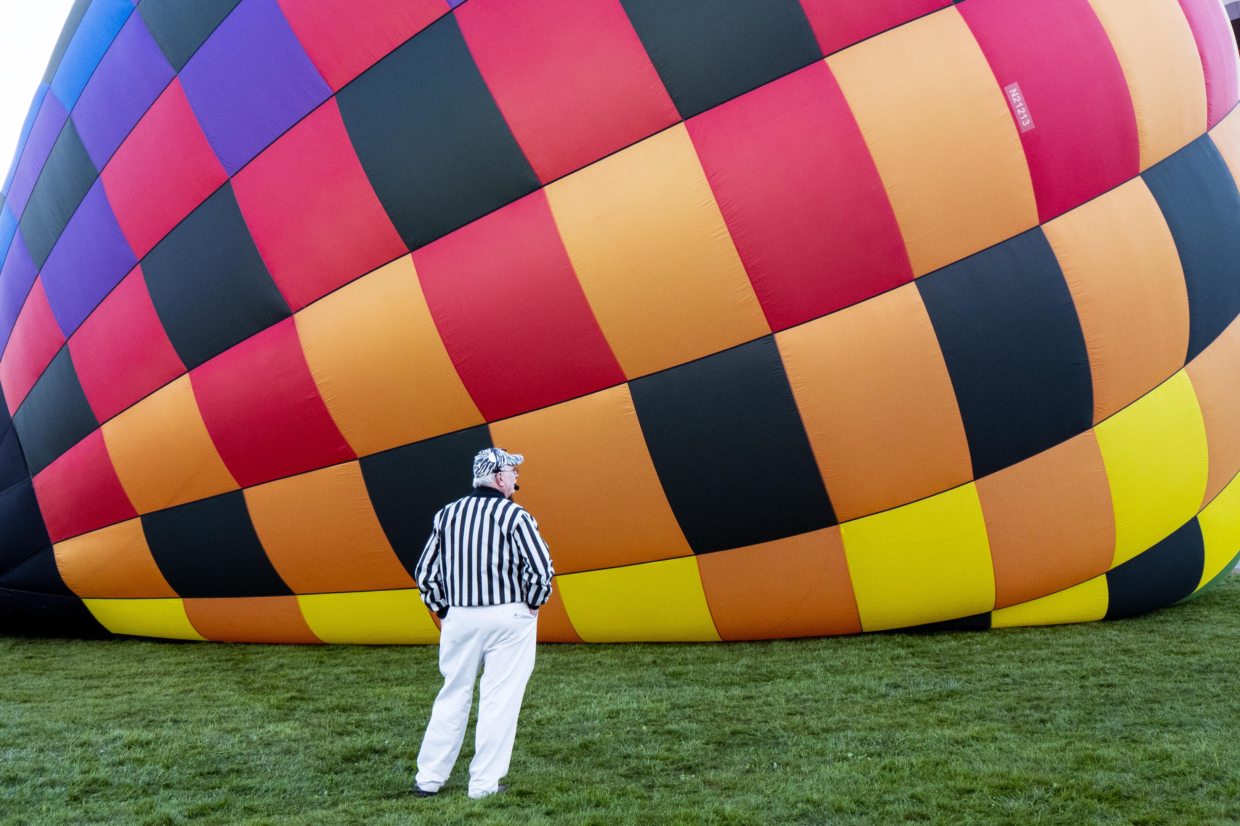 A launch coordinator wearing a black and white striped referee shirt watches a hot air balloon inflate on its side on the grass during the 2018 Albuquerque International Balloon Fiesta