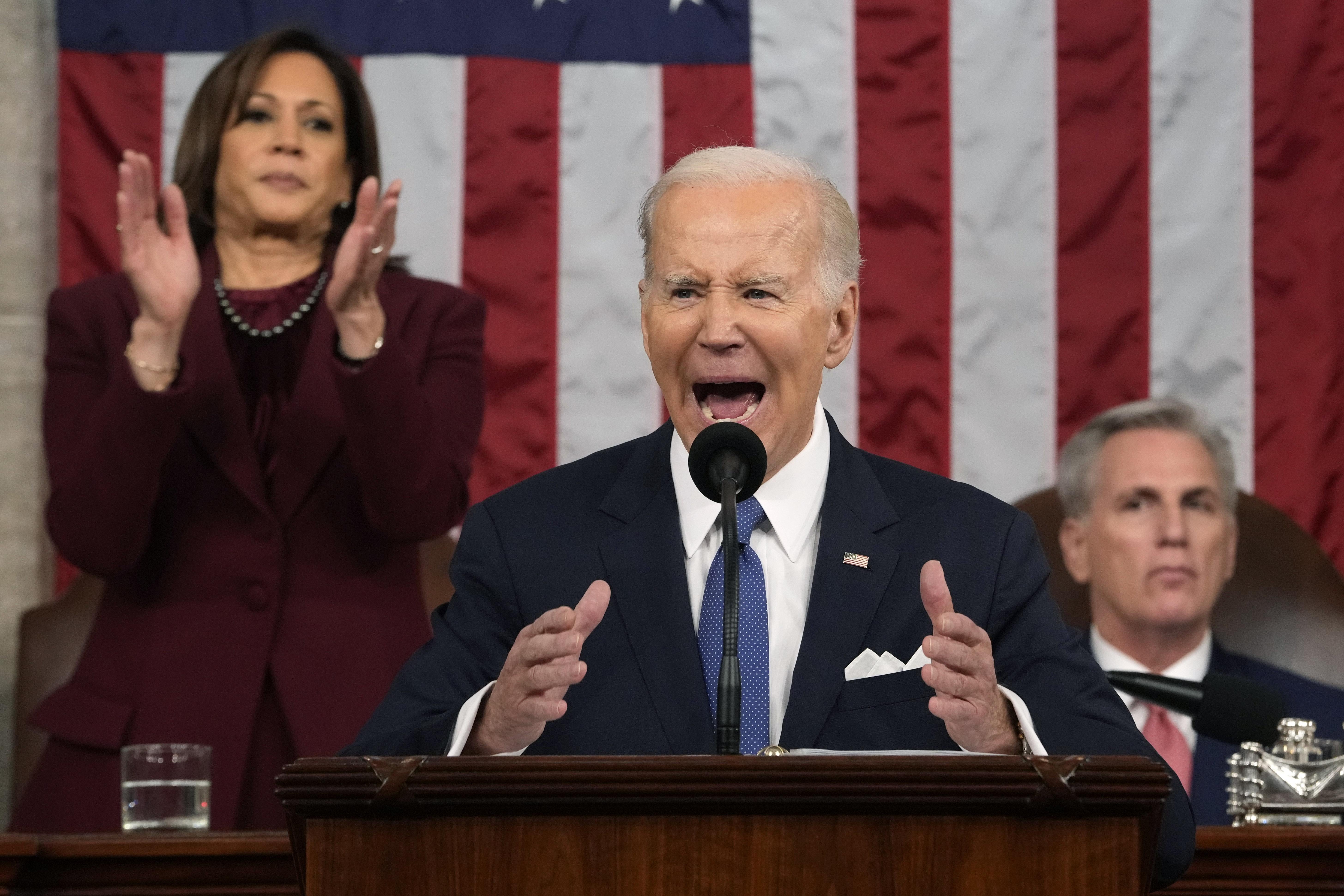 Joe Biden speaks into a microphone while Kamala Harris stands up to clap and Kevin McCarthy remains seated.